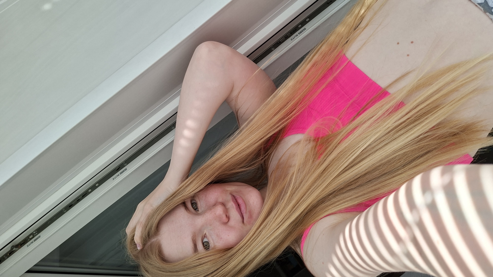 A woman with long, light blonde hair is positioned indoors, likely in Chaikovsky, Russia, as suggested by the image context. She's wearing a bright pink top. Her left arm is bent and her hand is in her hair. She is looking at the camera with a slight smile. She is near a window frame. Shadows from outside are cast on her arm.
