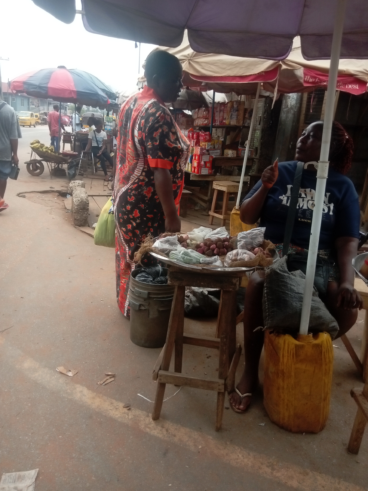 The image depicts an outdoor scene in Nkpor, Nigeria. A woman in a black and red patterned dress stands at a street-side stall. She is facing a second woman seated on a yellow plastic container, behind a table displaying produce, likely for sale. The seated woman is wearing a dark-colored t-shirt. Overhead, two umbrellas provide shade for the stall. In the background, other people are visible along with a wheelbarrow filled with yellow bananas. A building with a red and blue umbrella can be seen in the back. The street has a worn appearance.