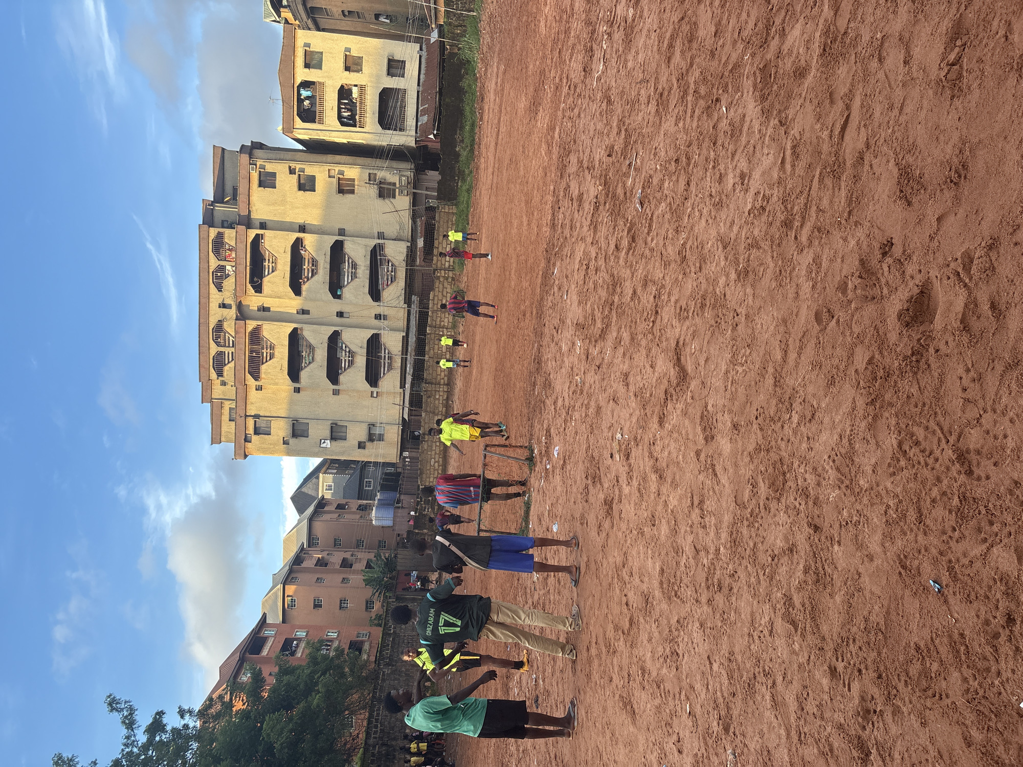 This is a photograph of a dirt soccer field in Onitsha, Nigeria, with a group of young men playing soccer. The field is made of reddish-brown dirt and is uneven, with some debris scattered across it. In the background, there are several multi-story buildings, some of which appear to be apartments or residential complexes. The sky is partly cloudy with blue sky visible.
On the field, there are at least ten individuals, most of whom are dressed in athletic wear, suggesting a soccer game or practice. Some are actively engaged in playing, with one person in a green shirt and black shorts on the left, and others scattered across the field in various colored shirts and shorts. One individual in the foreground wears a dark green shirt with the number "17" and the name "CHIZARAM" on the back. Another person in a striped red and blue shirt is visible near a makeshift soccer goal. The overall impression is a casual, community-based soccer game taking place in an urban environment.