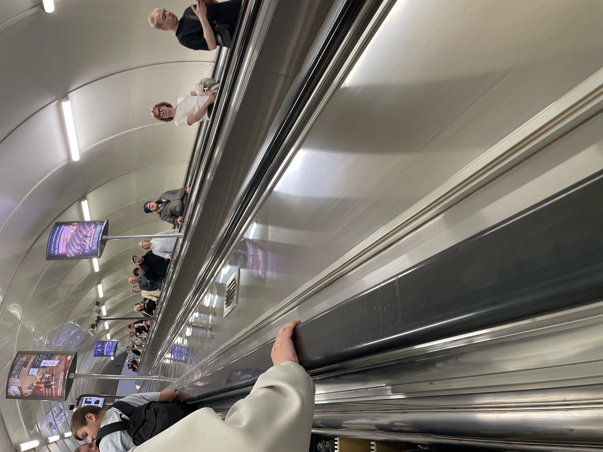 The image depicts an escalator in a subway station, likely in Saint Petersburg, Russia, given the presence of Cyrillic text on advertisements. Numerous people are ascending the escalator, some looking forward, others down at their phones. On the left, a person wearing a light-colored jacket and a backpack is visible. Further up the escalator, various individuals are seen in casual attire. Two prominent advertisements are mounted on the ceiling: one displays an illustration of people on skateboards, and another advertises a "BBQ Restaurant" with an image of grilled ribs. The escalator itself is made of polished metal, reflecting the overhead lights and the movement of the passengers. The station has a modern, curved ceiling structure with integrated lighting. The scene captures a moment of everyday transit within an urban environment.
