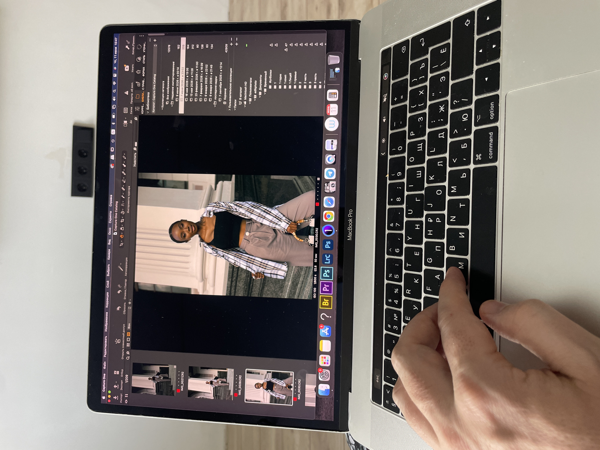 A person's hand is typing on a silver MacBook Pro. The laptop screen displays Capture One software, showing a photograph of a young Black woman in a plaid shirt and grey pants, standing on steps in front of a building with columns. The software interface includes file management options, image editing tools, and metadata for the displayed image. The environment appears to be indoors, with a portion of a wall visible in the background. The time on the laptop screen indicates 10:47 on June 11th. There is no visible indication of the city of Kazan, Russia in the image.