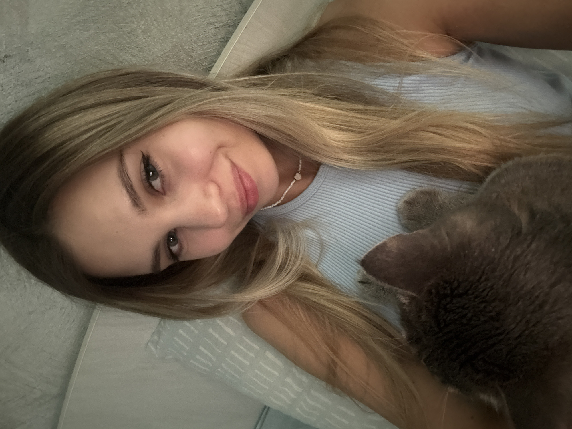 The image is a selfie of a young woman with blonde hair lying in bed, holding a grey cat. The woman is wearing a light blue ribbed tank top and a delicate beaded necklace with a small heart pendant. Her eyes are green, and she has light makeup on, including eyeliner. She is smiling faintly. The cat's head and part of its body are visible, nestled against her. The background consists of a light-colored patterned pillow and a textured wall or headboard. There is no indication of the city of Chaikovsky, Russia.