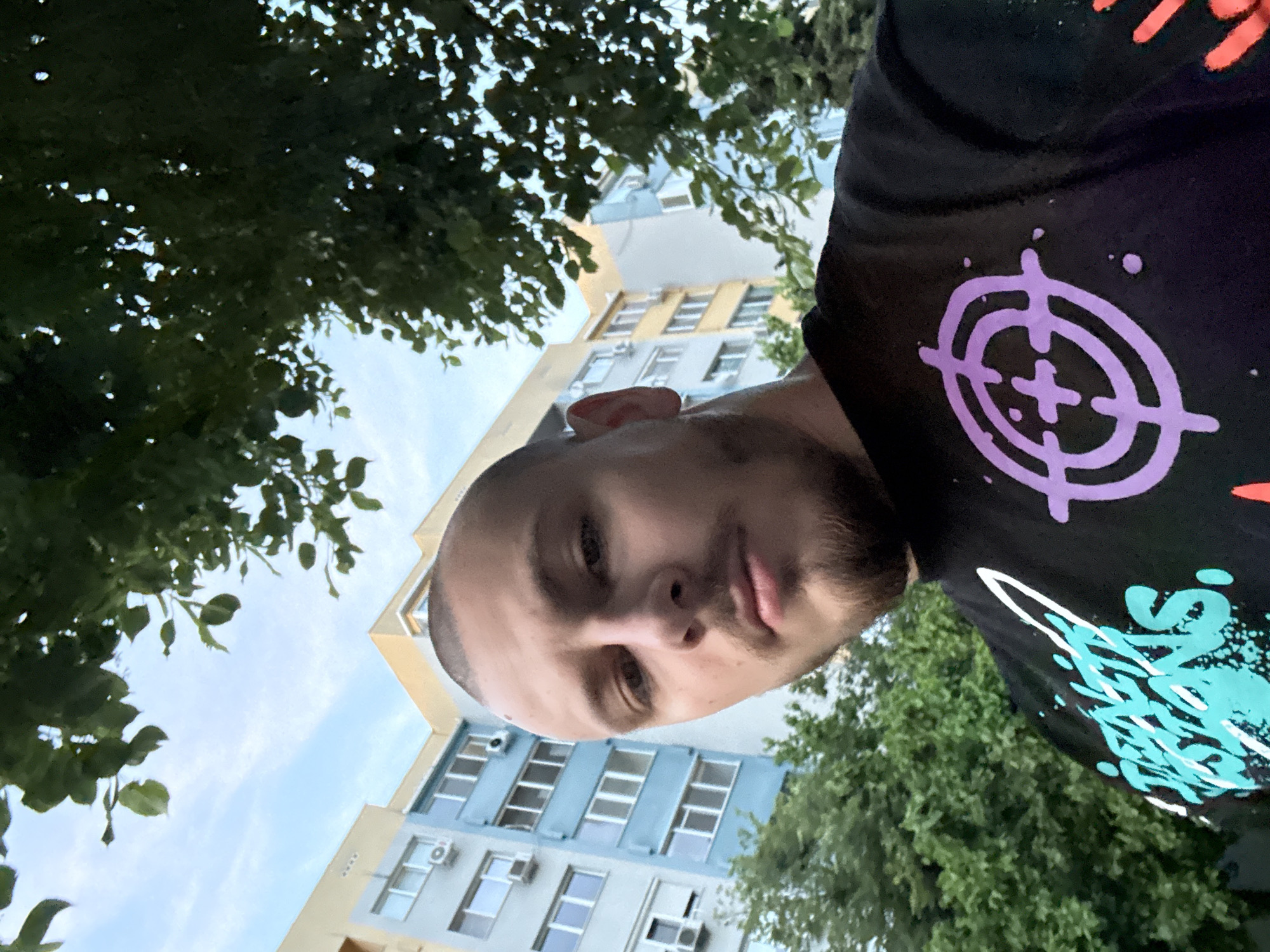 A selfie taken outdoors features a man in the foreground wearing a black T-shirt with a purple target symbol and teal and white graphics. He has short, dark hair and a trimmed beard. The background includes a building with a yellow top and blue-grey lower floors with white-framed windows. Air conditioning units are attached below some of the windows. Green trees are partially visible on either side of the frame. The sky is light blue, indicating daylight. The location could possibly be Stara Zagora, Bulgaria, though no details in the photograph conclusively confirm this.