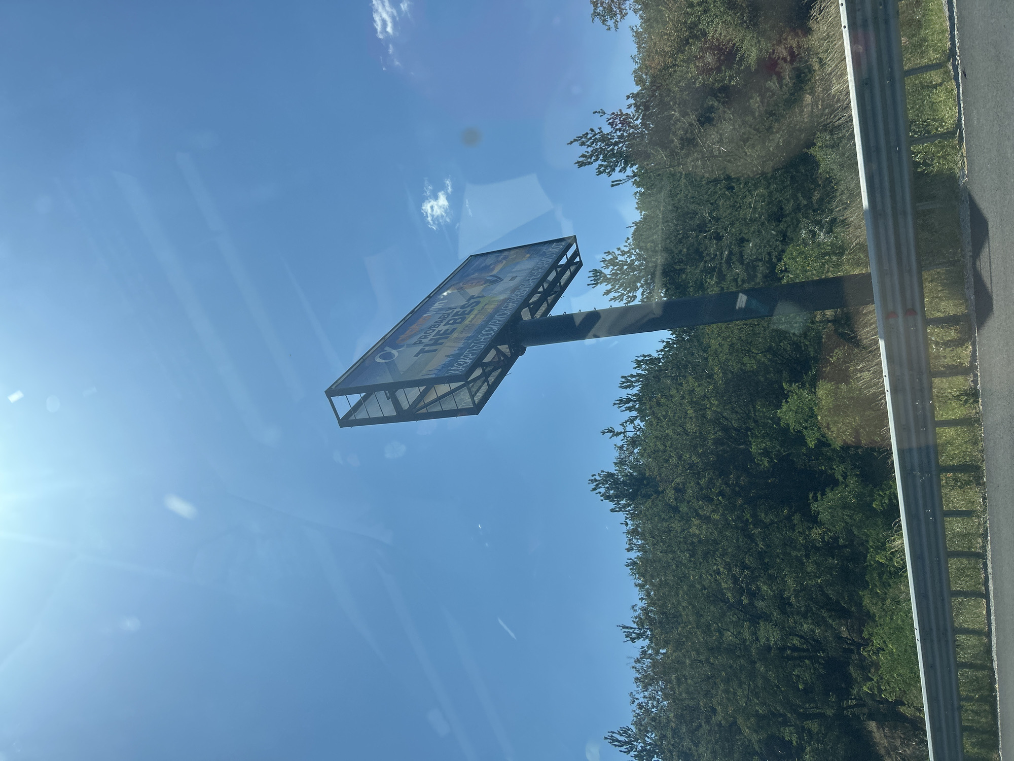 A low-angle, medium shot captures a large billboard situated in a rural setting, with a bright sun glaring down from the upper left corner, casting lens flares and obscuring details of the blue sky. The billboard, mounted on a dark metal pole, displays an advertisement with prominent text reading "PROBABLY THE BEST" in a large, white, sans-serif font. Below this, a line of text in a smaller, less legible font is visible. The billboard's content appears to be a vibrant advertisement featuring two individuals, one in a suit and another in casual attire, suggesting a promotional campaign.
The billboard stands against a backdrop of a dense, green forest with varying shades of green indicating different types of foliage and tree heights. The forest line extends across the middle ground of the image. In the foreground, a metal guardrail, typical of roadside infrastructure, runs horizontally, separating the viewer's perspective from the grassy area and the dark asphalt of a road. The overall environment suggests a roadside location, likely along a highway or main road in a rural or semi-rural area. The specific location in Vakarel, Bulgaria, is provided as context, though it is not visually discernible from the image alone. The day is sunny and clear, with a few small, wispy clouds visible in the sky.
