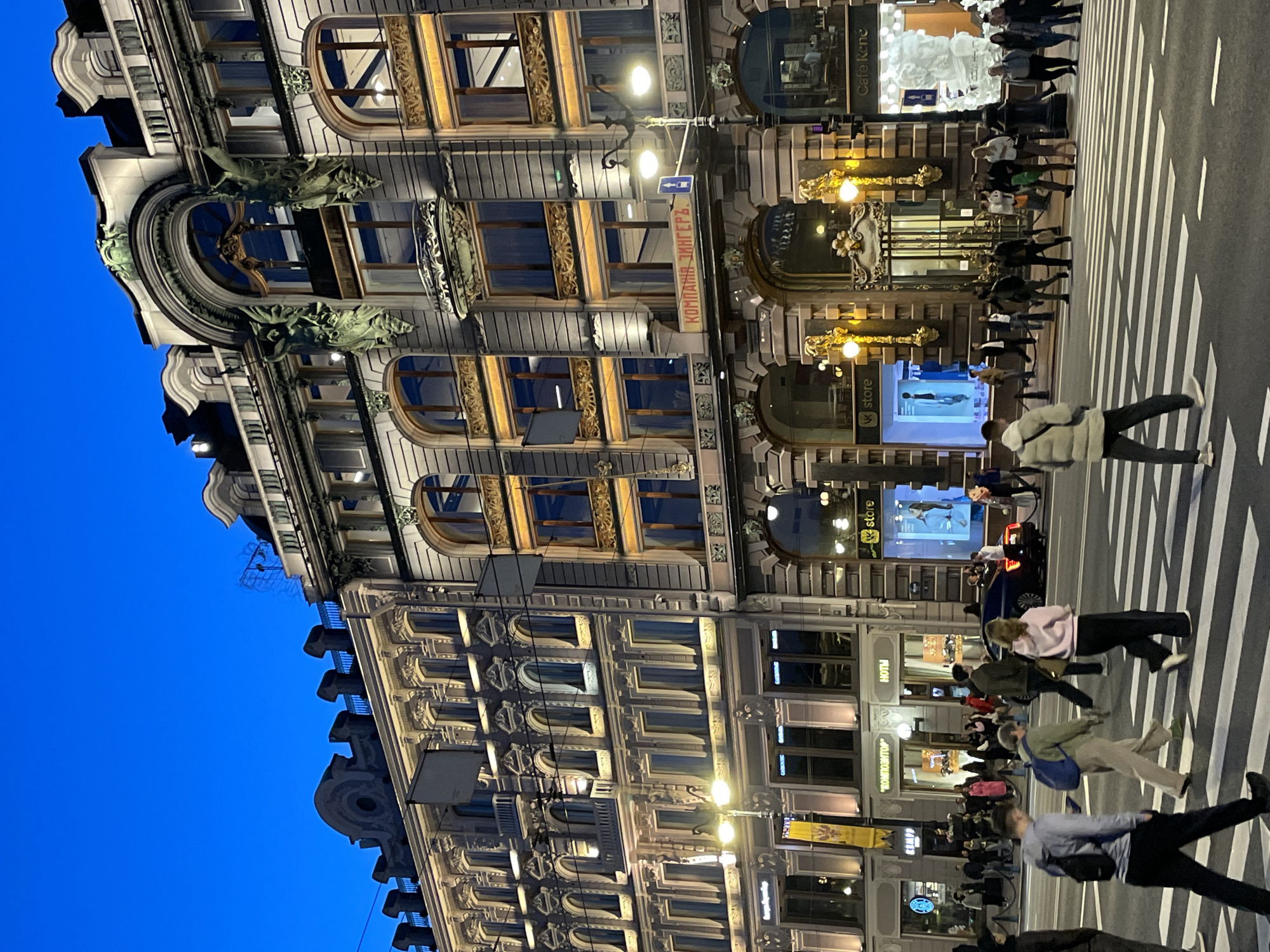 The image captures a vibrant urban street scene at dusk or early night in Saint Petersburg, Russia. A grand, ornate multi-story building dominates the left side of the frame, its detailed facade illuminated by warm yellow lights. This historic structure features a blend of architectural styles, including rounded windows, decorative carvings, balcony railings, and sculptural elements resembling gargoyles or mythical creatures above some windows.
On a prominent sign on the building, the Cyrillic text "КОМПАНИЯ ЗИНГЕРЪ" (Kompania Zinger, or Singer Company) is clearly visible, identifying the building as the iconic Singer House, now known as Dom Knigi (House of Books). Other visible text includes "Cafe Rene" and "vk store" on storefronts across the street, as well as "HOMN", "БАНКРОТСТВО" (BANKROTSTVO), and "КНИГА" (KNIGA, or book) on various signs. A blue traffic sign with an arrow points left.
The street in the foreground and to the right is bustling with activity. Numerous pedestrians are seen walking, many crossing a wide zebra-striped crosswalk. Their movement suggests a typical evening in a busy city center. Streetlights cast bright circles of light, further illuminating the scene. The sky above is a deep, clear blue, transitioning from twilight to night. A few vehicles are visible in the background, but the focus remains on the pedestrian activity and the illuminated architecture. The overall impression is one of a lively, historically rich urban environment.