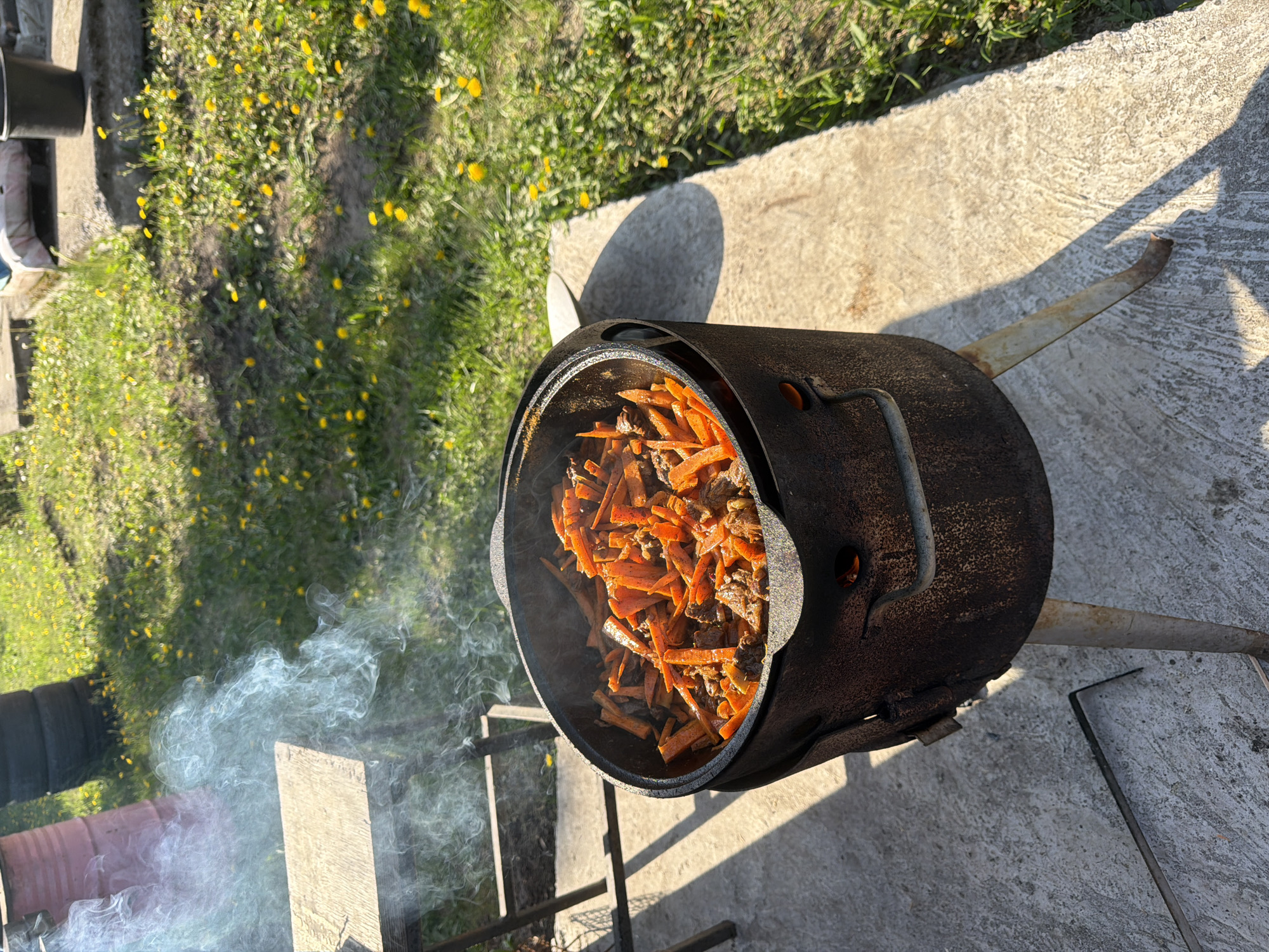 The image depicts a cooking pot filled with what appears to be stewed meat and sliced carrots. Smoke is emanating from the cooking area. The pot is placed on a metallic, tripod-like stand, suggesting outdoor cooking. The scene is set on a concrete surface, with grass and dandelions visible in the background. The location is identified as Большая Шильна, Russia.
