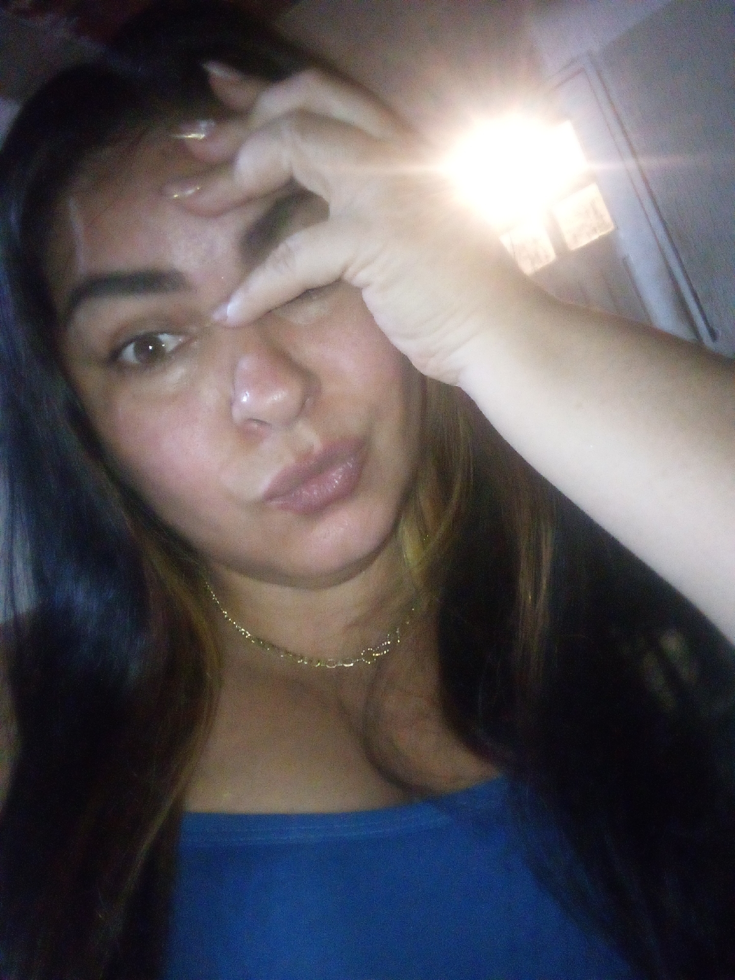 The image depicts an adult female posing for a close-up selfie indoors, likely within a private residence in Juan Griego, Venezuela. The subject occupies the majority of the frame, positioned from the chest up. She has long, dark hair with visible lighter strands, dark eyes, and medium skin tone. Her left hand is raised, partially obscuring her left eye and forehead, with her fingers slightly parted. Her right eye is open and directed towards the camera. Her lips are slightly pursed. She wears a plain blue strapless top and a thin gold-colored chain necklace. A bright light source, possibly a camera flash or an overhead fixture, is prominent in the upper right background, causing significant glare and overexposure. Behind the subject, a light-colored wall or door frame is partially visible, featuring indistinct rectangular elements near the light source. The overall environment appears to be an interior room.