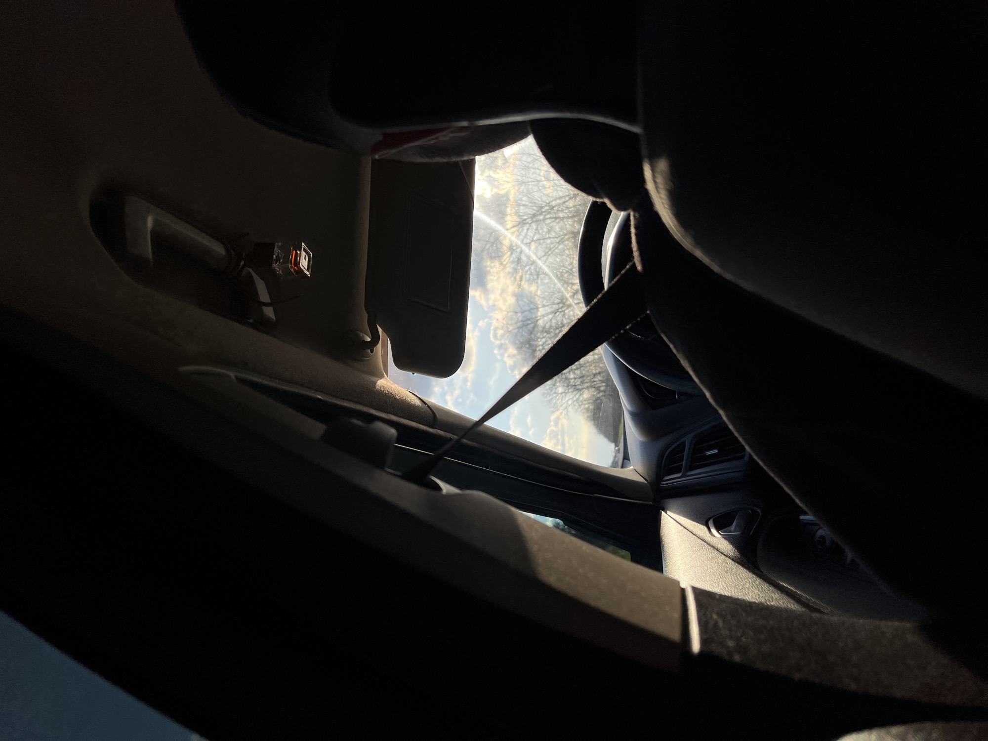 The image captures an interior view from a car, looking out through the front windshield and the passenger-side window. The scene is strongly backlit by a bright sun, creating prominent glare and rendering much of the car's interior in silhouette.
On the left, the car's headliner and the underside of the passenger sun visor, which is in an upward position, are visible. A small, rectangular device with an orange glow, possibly a sensor or interior light, is affixed to the headliner, secured with what appear to be zip ties. A seatbelt strap extends diagonally from the upper right, crossing the middle of the frame.
Towards the right, portions of the dashboard, including air vents, and part of a steering wheel are discernible, primarily as dark shapes against the bright exterior.
Outside the vehicle, a bright sky with scattered clouds dominates the upper half of the view. The sun is clearly visible, producing significant lens flare. Below the sky, silhouetted bare tree branches suggest a winter or early spring setting. A distant view of the ground or road is minimally visible.
The intense, low-angle sunlight indicates either early morning or late afternoon. The weather appears clear with some cloud cover. No specific activity or human subjects are visible. There is no discernible text within the image. The location context is Ternovskaya, Russia.
