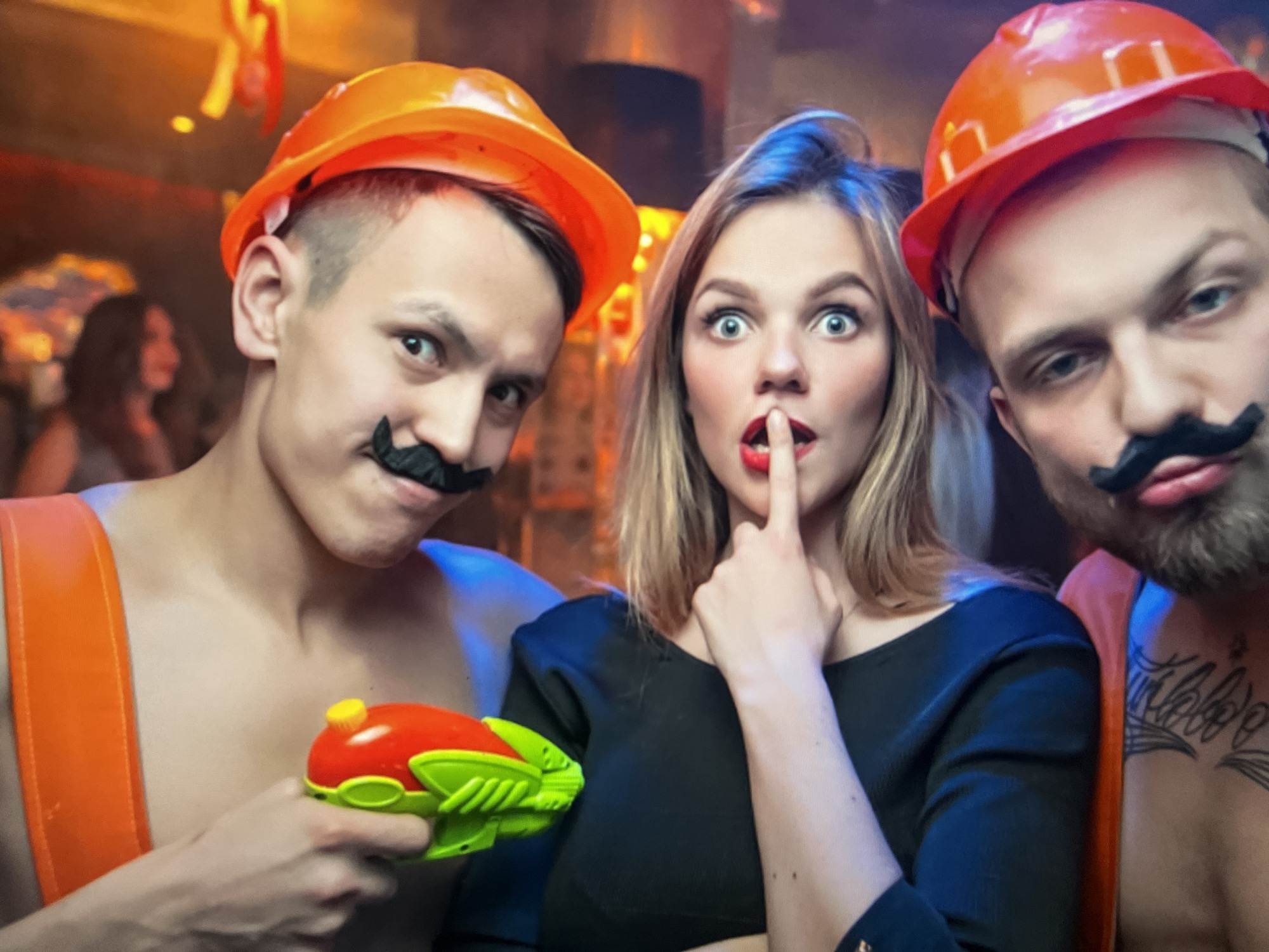 Three people are posing for a photo. Two men, wearing orange hard hats and fake mustaches, are on either side of a woman. The woman is wearing a black top and has her finger to her lips, as if telling a secret. The man on the left is holding a red and green water gun. The background appears to be a dimly lit indoor venue, possibly a bar or club, with decorations and other people visible in the background. The location is not identifiable as Qarağandı city administration, Kazakhstan.