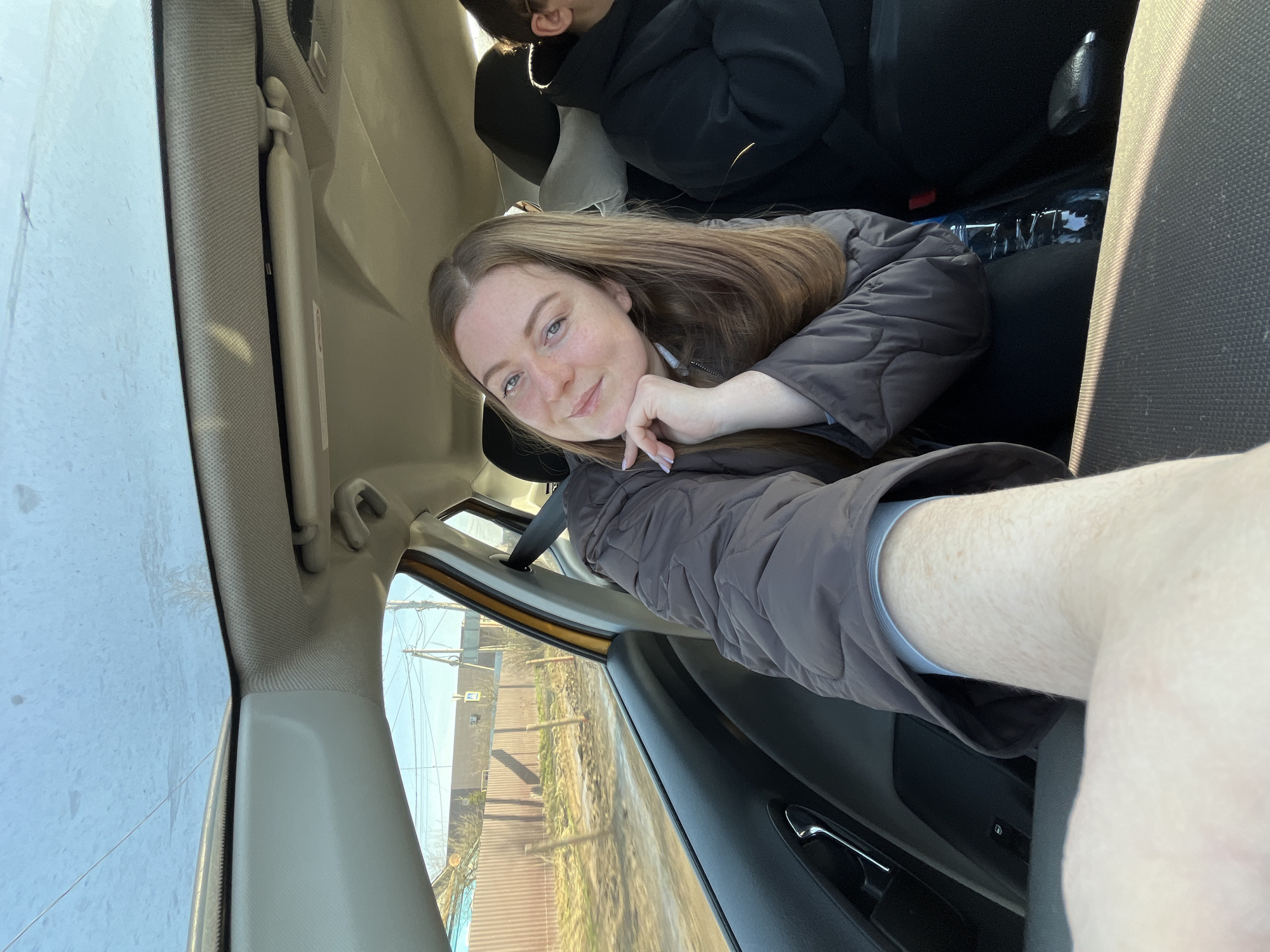 A woman is in a car. She is facing the camera, resting her head on her hand. She is wearing a brown jacket. There is another person in the back seat of the car. The car is moving past a street. It is a sunny day. The location is likely Yasenevo, Russia, based on visual features in the background such as building design and foliage.