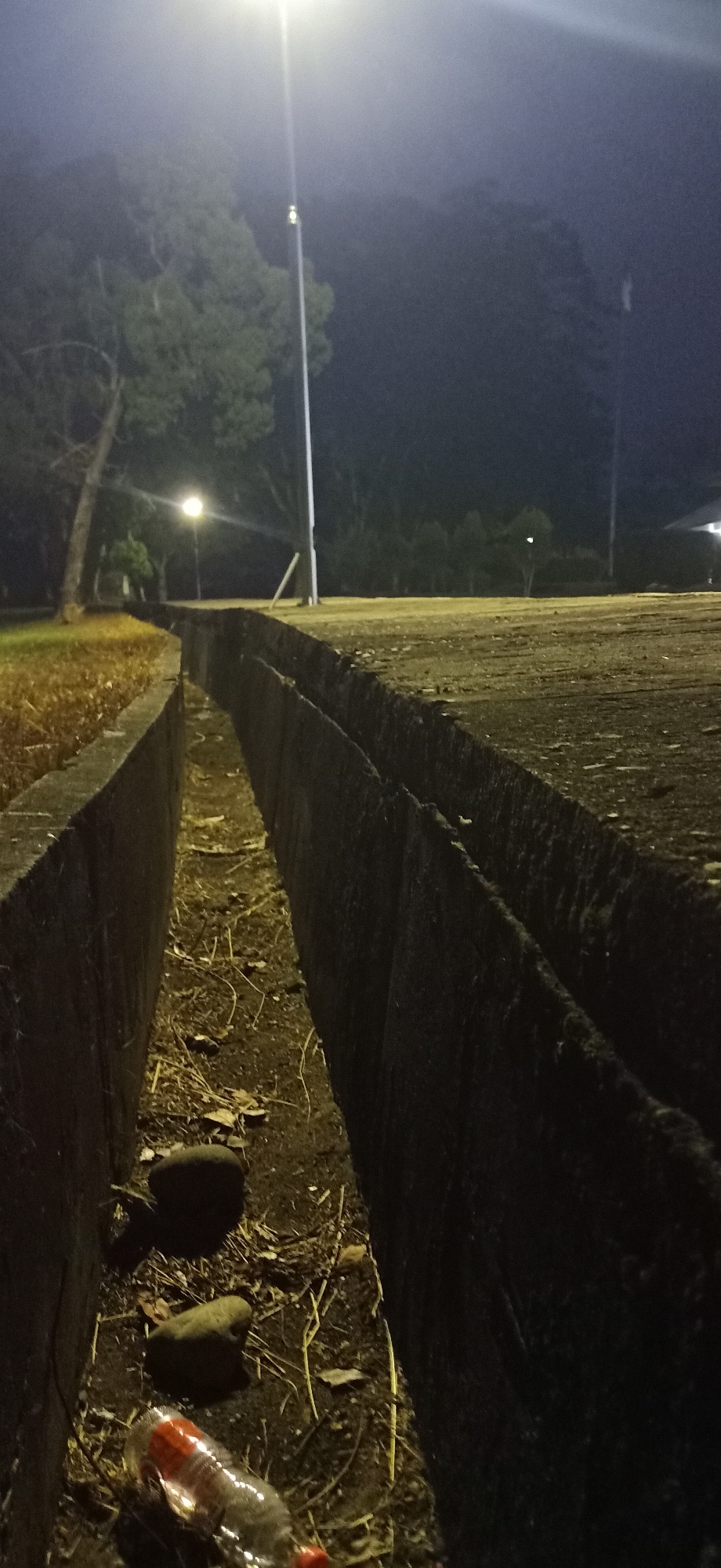 The image depicts a drainage ditch at night in Palohgadeng, Indonesia, illuminated by streetlights. The ditch is made of concrete and runs along a paved surface, possibly a park or walkway. The bottom of the ditch contains dirt, dry leaves, twigs, and two stones. A partially visible plastic bottle with a red cap lies on its side in the ditch. In the background, trees and a tall pole with a light fixture are visible, along with a misty atmosphere. The overall impression is of a somewhat neglected outdoor area at night.
