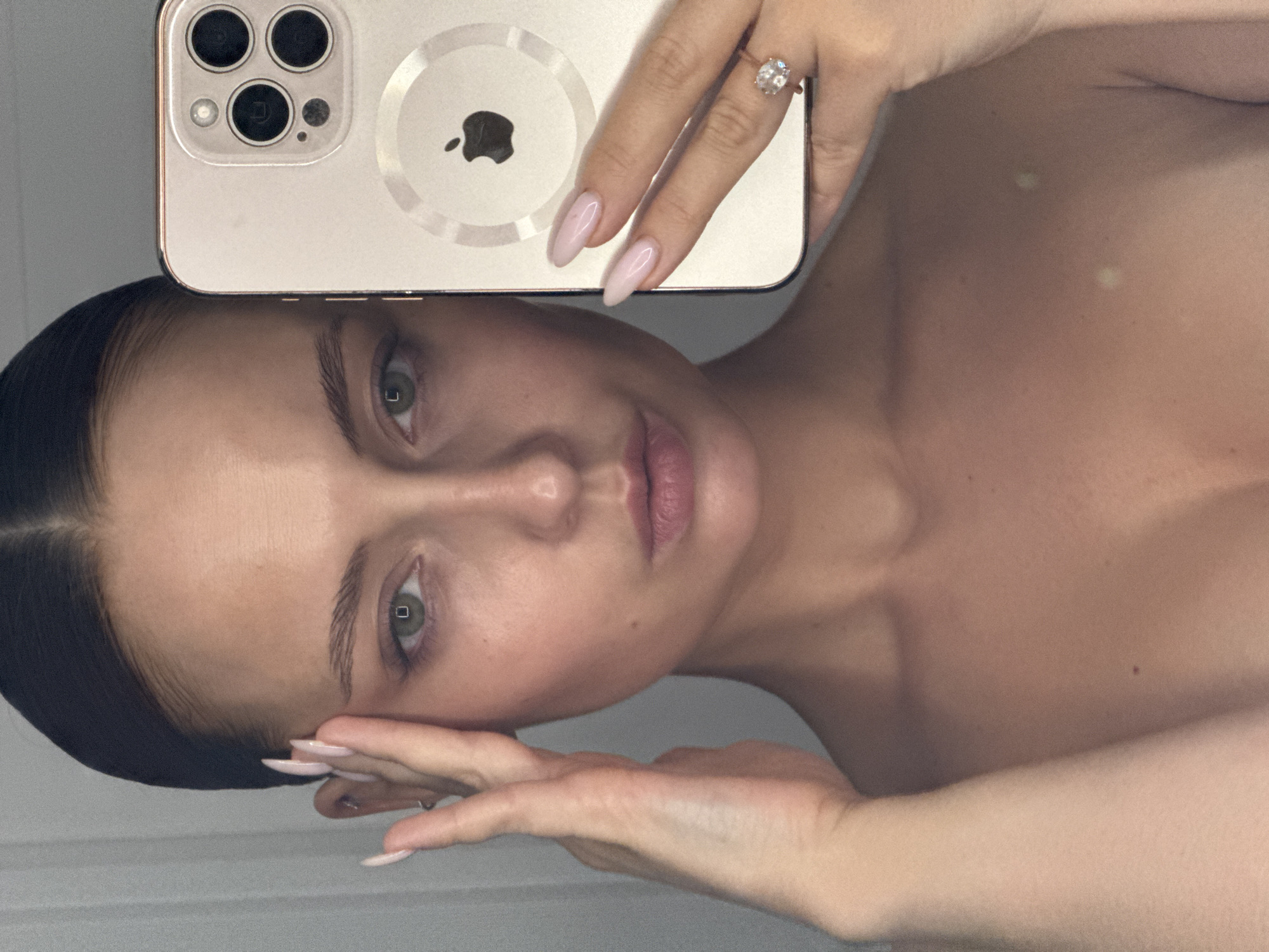A woman with dark hair slicked back takes a selfie with a champagne-colored iPhone. The phone partially obscures her forehead. She holds the phone with her left hand, displaying an oval-cut diamond ring and pink manicured nails. Her right hand is near her face, also displaying pink manicured nails. Her expression is neutral. The background is a plain, light-colored wall. There is no indication of the location being in Chaikovsky, Russia.