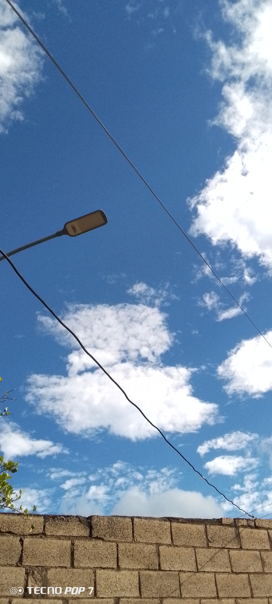 The image shows a partly cloudy, blue sky above a brick wall and a street light in Altagracia, Venezuela. Two wires are visible running across the sky. The brick wall is in the foreground, and some greenery is visible on the left. "Tecno Pop 7" is displayed in the bottom left corner, likely indicating the device used to take the picture.