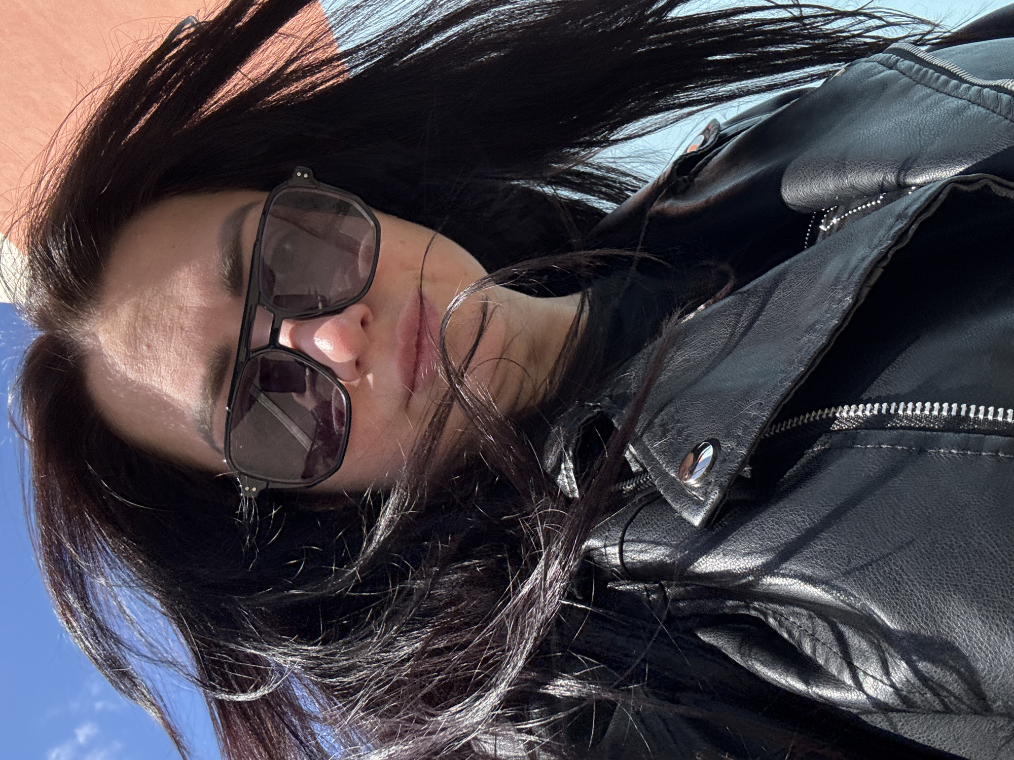 The image is an angled, close-up portrait of a person, likely a woman, with long, dark, straight hair and wearing sunglasses. The subject's head is tilted, and their body is positioned diagonally across the frame. Their dark hair is swept upwards and to the left, appearing to be in motion or styled to one side.
The person wears large, black-framed sunglasses with dark lenses, obscuring their eyes. A slight, relaxed expression is visible around their mouth. They are dressed in a black leather jacket, detailed with a prominent silver-toned zipper and a metallic snap button on the collar/lapel, visible on the right side of the frame. The texture of the leather is evident due to the bright, direct sunlight.
The setting is outdoors during the day, indicated by the bright, clear blue sky visible in the background, particularly in the lower left and right sections. A light-colored, possibly pale orange or pinkish, surface is visible in the upper left background. The strong sunlight creates distinct highlights on the hair, sunglasses, and jacket, suggesting sunny weather. No visible text is present in the image. The overall impression is casual and confident.