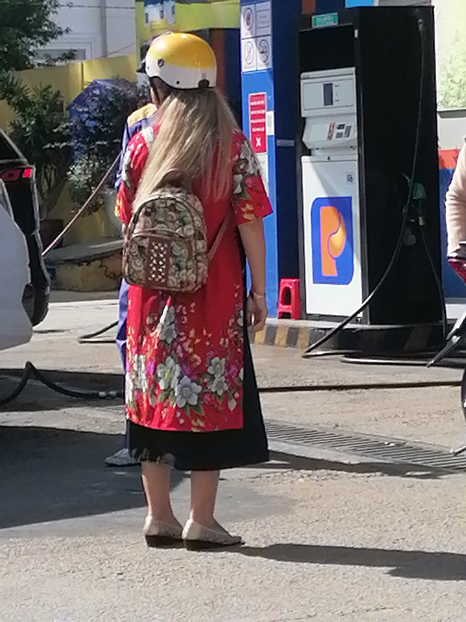 The image depicts a woman standing at a gas station in Da Lat, Vietnam. She is wearing a yellow helmet, a red floral patterned dress, and a black skirt. A patterned backpack is slung over her shoulders. She is standing on a paved surface with yellow and black striped markings. In the background, there is a blue and black gas pump with a blue and orange logo. Other individuals and objects, including a car and a motorcycle, are partially visible. The scene is outdoors, with sunlight casting shadows on the ground.