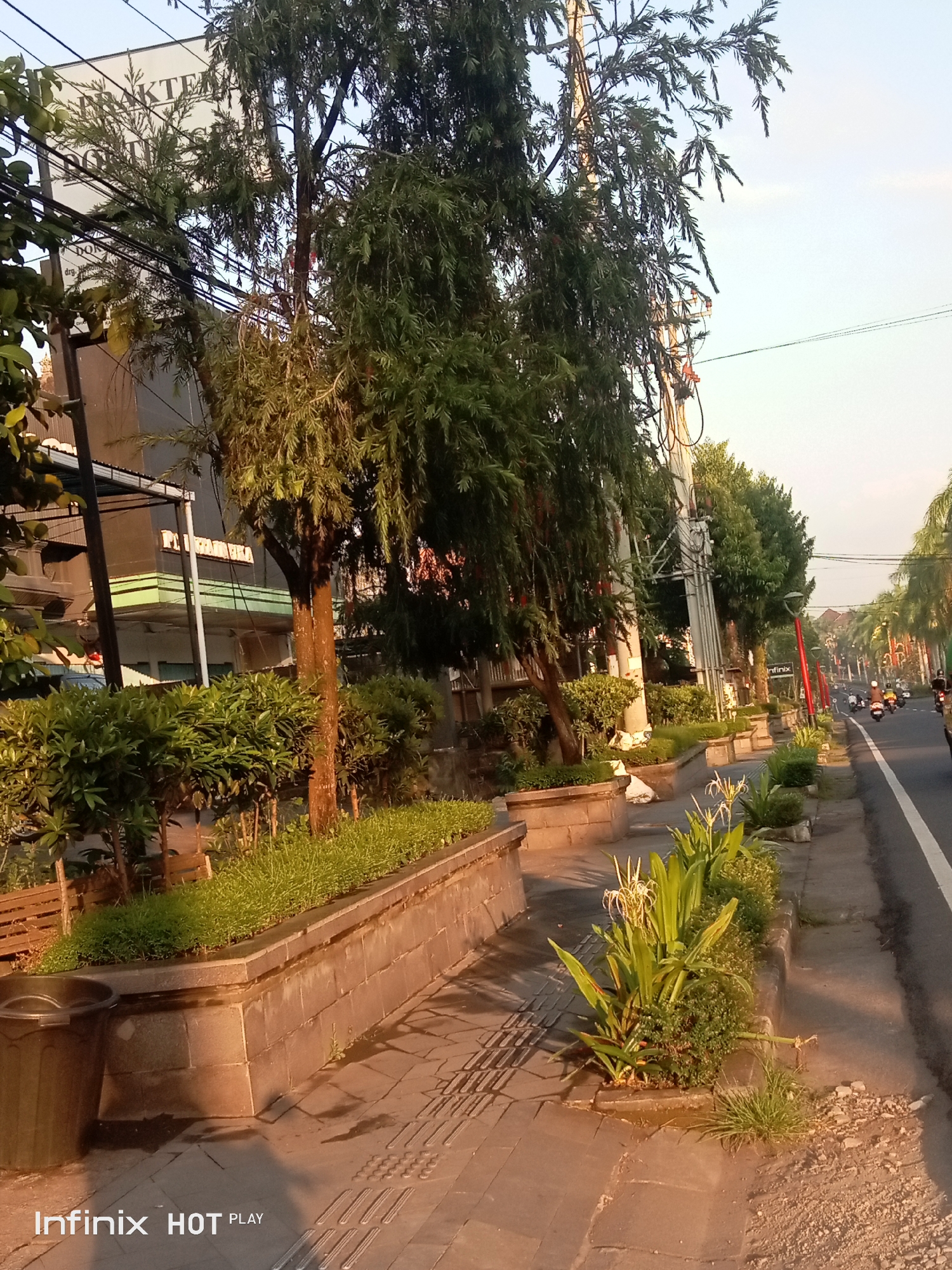 This image depicts a street scene in Gianyar, Indonesia, during what appears to be late afternoon or early evening, suggested by the warm, low-angle lighting. The foreground features a paved sidewalk with textured tactile paving, indicating a pedestrian area. A dark, large trash bin stands on the left. Adjacent to the sidewalk is a raised planter box constructed of stone blocks, filled with green foliage and plants, including some flowering plants with white, spidery blossoms.
Further along the sidewalk, more planter boxes and trees line the street. Several power poles with numerous wires stretch across the scene. Across the street, there is a road with a white lane marker, where several motorcycles are visible, some with riders. The buildings on the opposite side of the street are partially obscured by trees and the angle of the shot, but one building displays a sign with Indonesian text, possibly indicating a business or clinic. The overall impression is that of a well-maintained urban street with a focus on green spaces. The Infinix HOT PLAY watermark is present in the bottom left corner, indicating the device used for capture.