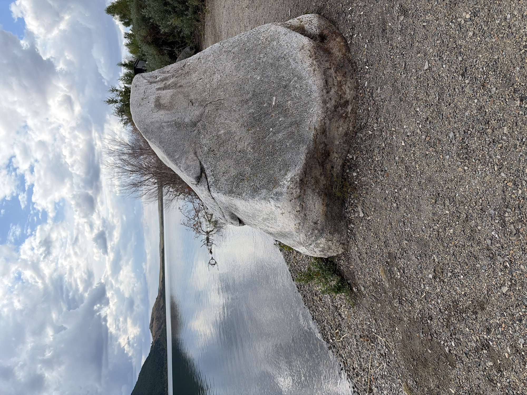 The image depicts a large, grey boulder situated on a gravelly shore next to a body of water. The water reflects the cloudy sky and a distant, tree-covered hillside. In the background, there are sparse trees and a small structure, possibly a shed or cabin. The ground is covered in small, mixed-colored pebbles. The sky is filled with dramatic, grey clouds with patches of blue sky visible. The scene appears to be in a natural, outdoor setting, possibly near a lake or reservoir.