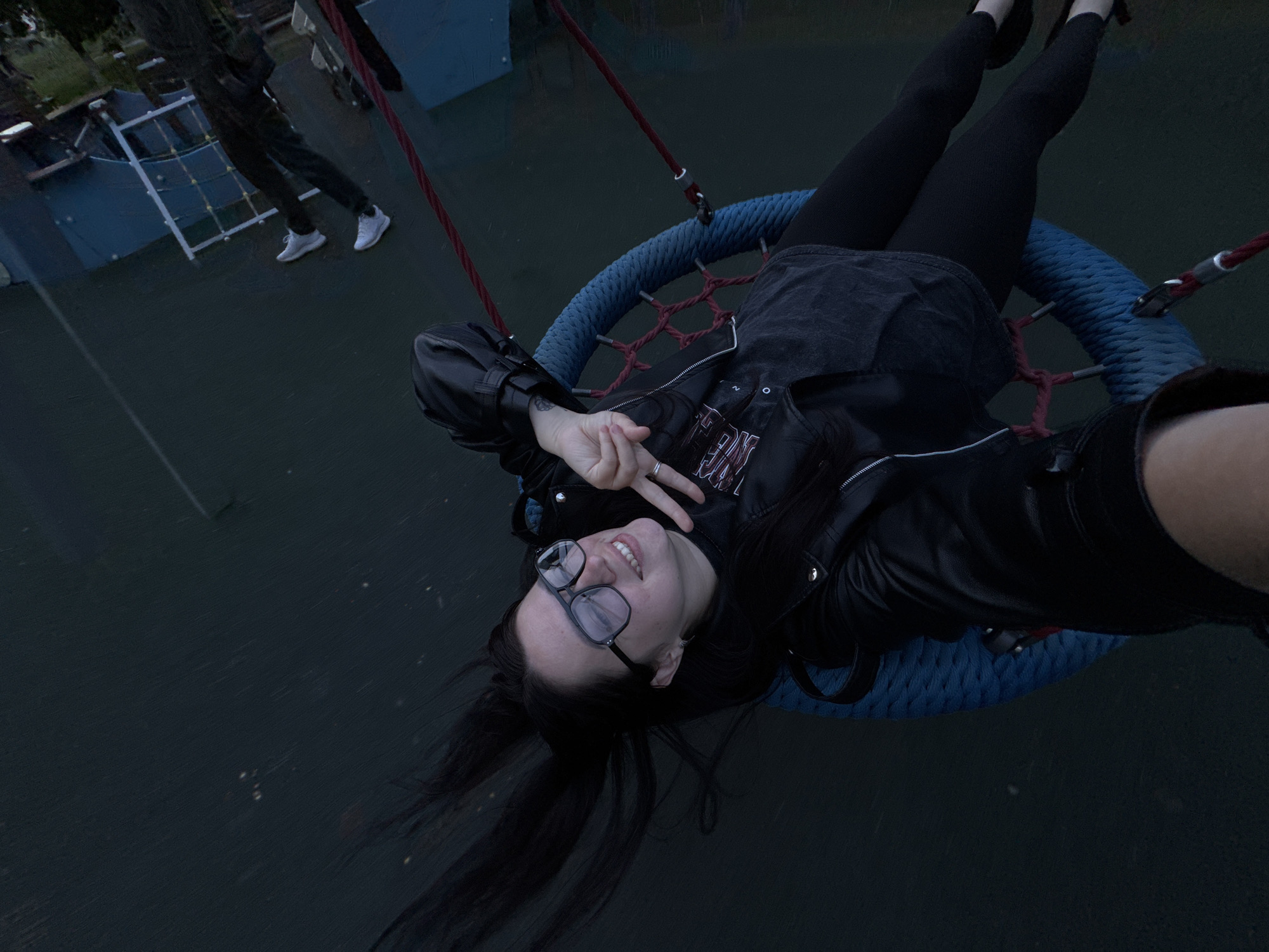 An overhead shot captures a young woman lying on her back on a large, round blue and red net swing. She is smiling broadly, wearing dark-framed glasses, and holding up two fingers in a peace sign towards the camera, which she appears to be holding herself for a selfie. Her long, dark hair is spread out behind her. She is dressed in a black leather jacket over a dark top that has some partially visible white lettering (possibly "NGE"). Her legs are bent at the knees, and she wears black leggings.
In the blurred background, another person, dressed in dark clothing and white sneakers, stands near a playground structure with white frames and blue panels. The scene appears to be set outdoors in a park or playground. The lighting suggests it is either dusk or nighttime, contributing to the dark, muted tones of the image. The ground beneath the swing and figures is dark. The overall mood is relaxed and cheerful, indicated by the woman's smile.