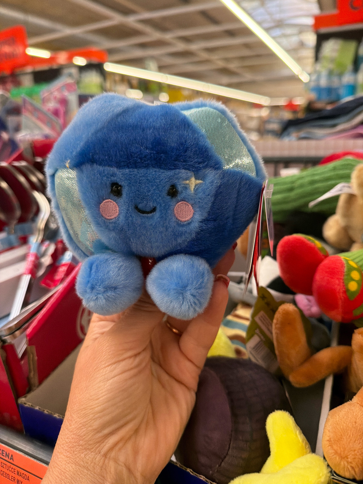 In Wrocław, Poland, a hand holds a small, round, blue plush toy with a smiling face, pink cheeks, and sparkling star accents. The toy has two puffy blue feet. The background appears to be a retail store aisle, with shelves stocked with various items, including more plush toys and clothing. Fluorescent lighting illuminates the scene, casting a slight glare.