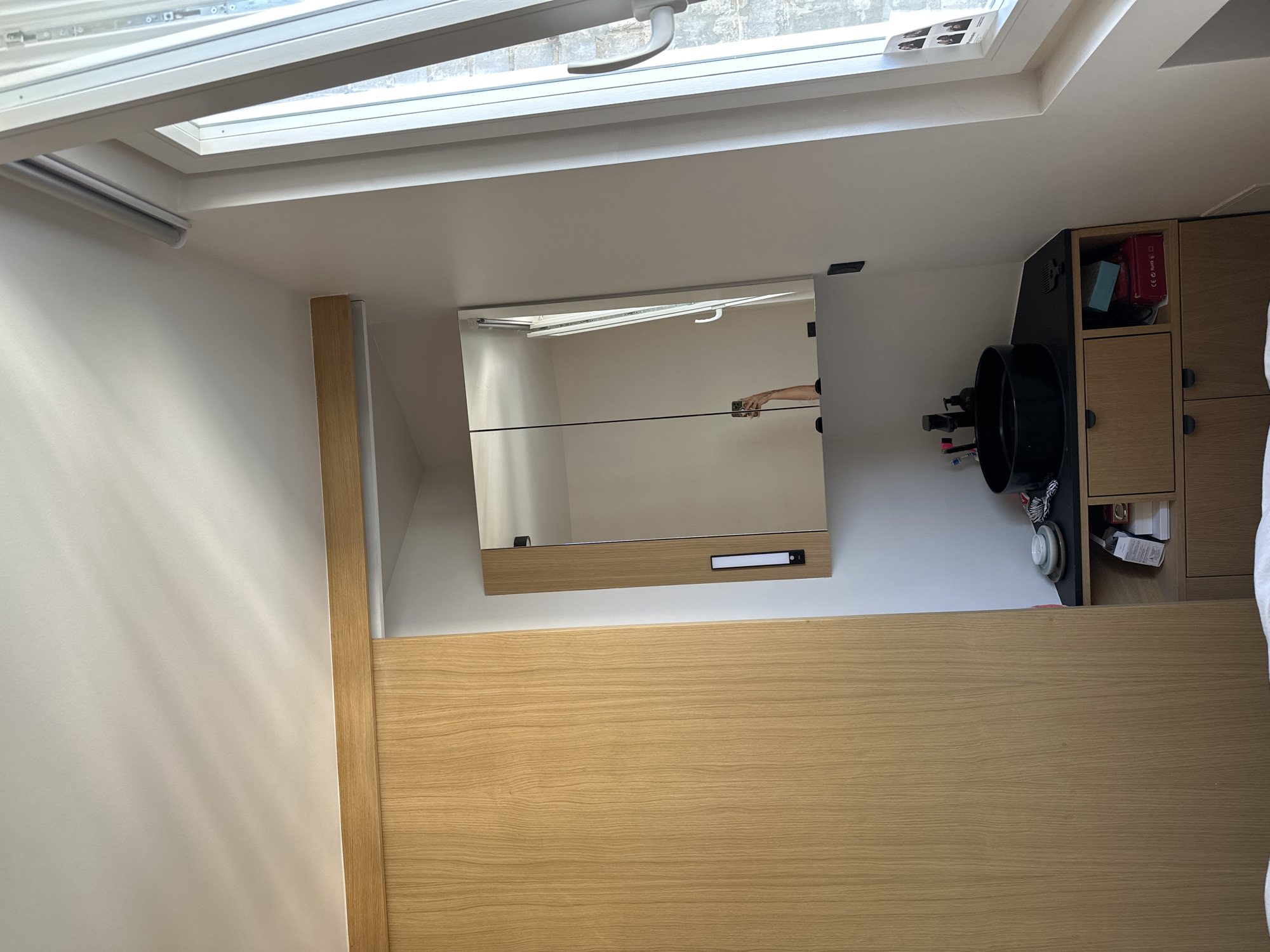 This appears to be an interior shot of a room, possibly a bathroom, with a modern aesthetic. The location is Saint-Ouen-sur-Seine, France.
The image is taken at an angle, showing a portion of the ceiling with a skylight. The skylight frame is white, and a window is visible.
Below the skylight, there is a white wall that connects to a wooden paneled wall. There is a mirror mounted on the wall. A reflection of a hand can be seen in the mirror.
Adjacent to the mirror and wooden paneling, a cabinet with drawers and shelves can be seen. There are items stored on the shelves and a black sink is to the left of the cabinets.