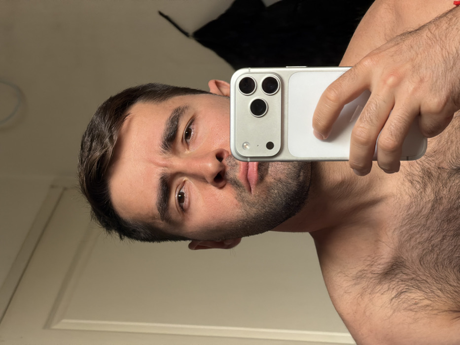 A Caucasian male, approximately 25-35 years old, with short dark brown hair, a dark stubble beard, and brown eyes, is depicted from the mid-chest upwards, shirtless, in an indoor setting within Tel-Aviv, Israel. He holds a light-colored smartphone, likely silver or white, in his right hand, positioning it to capture a mirror selfie. The phone features a prominent camera module with three dark circular lenses and a smaller circular element, and a light-colored accessory is attached to its back. The man has a slight pout and furrowed brow, looking directly towards the phone's lens/viewer. His chest displays visible dark hair. The background consists of a plain light-colored wall. A white door frame is partially visible on the left side of the frame. On the right, a dark, indistinct object hangs against the wall, with a faint light blue circular mark visible on the wall above it. The scene is illuminated by ambient indoor light.