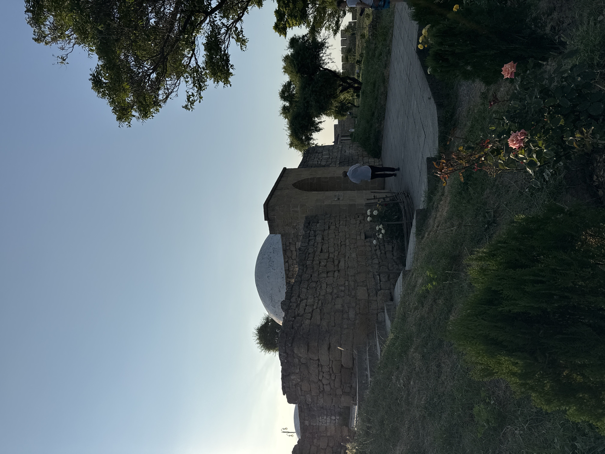A person walks away from the camera towards an arched gateway in a stone wall. The gateway leads into a courtyard area within a historical complex. The wall is constructed of rough-hewn stones and is topped by what appears to be battlements in the background. To the left of the gateway, a large, light-colored dome is visible, rising above the stone wall. Green trees and vegetation are present throughout the scene, with some flowering plants in the foreground. The sky is clear and blue, suggesting it is daytime. The overall setting suggests an ancient fortress or citadel.