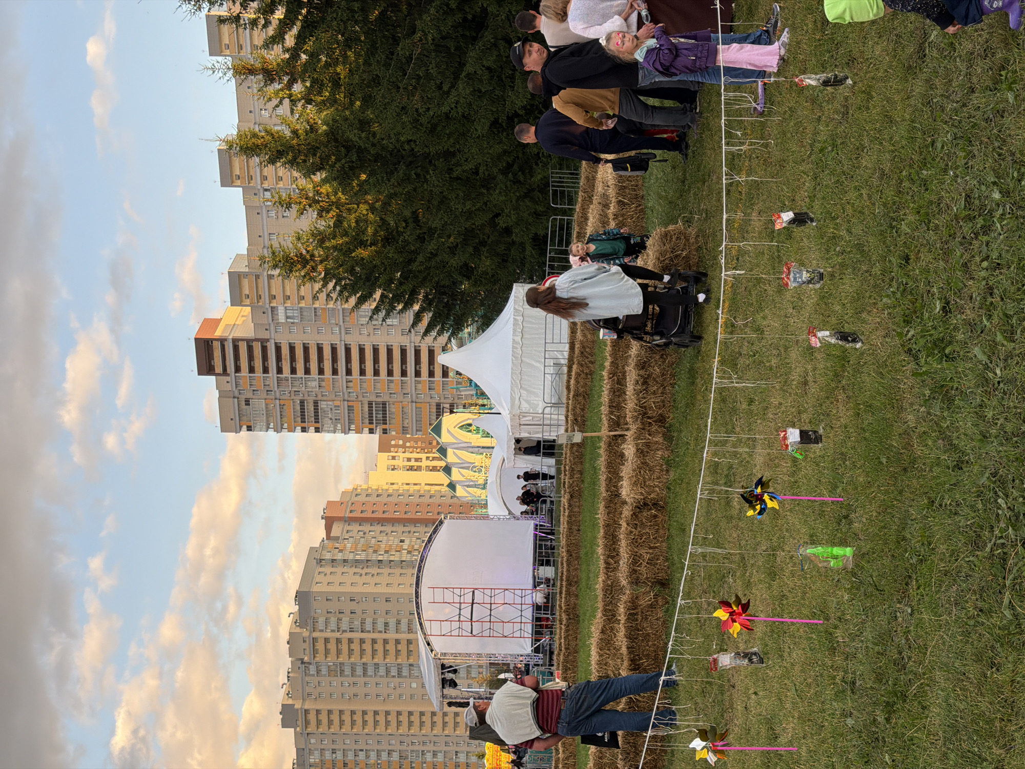 An outdoor event is taking place in an urban park. In the background, tall residential buildings rise against a cloudy sky. In the middle ground, a stage with a white screen is set up, and several white tents are present. A row of hay bales is laid out on the grass, suggesting a seating area or an activity space. People are scattered throughout the scene, some standing and observing, while others are walking. A woman is pushing a stroller, and a child is standing near her. In the foreground, a string of colorful pinwheels and small bagged items are hanging, likely part of a game or decoration. The ground is covered in green grass. The overall impression is of a community gathering or festival.