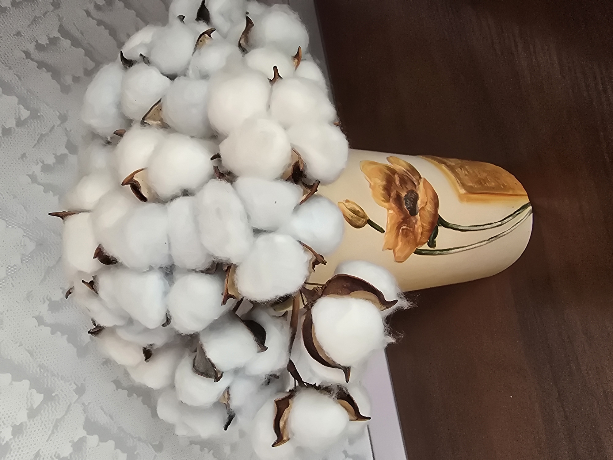 An arrangement of fluffy cotton bolls sits in a decorative vase. The vase is cream-colored and features a painted illustration of an orange poppy with a dark center and green stems. The vase itself has a textured, possibly metallic, square design in a golden hue on its side. The cotton bolls are predominantly white and appear to have brown remnants of the cotton plant stems attached. The background consists of a textured white curtain and a dark brown wooden surface. The lighting is soft, creating a gentle illumination of the subjects. This scene is located in Sofiivska Borshchahivka, Ukraine.