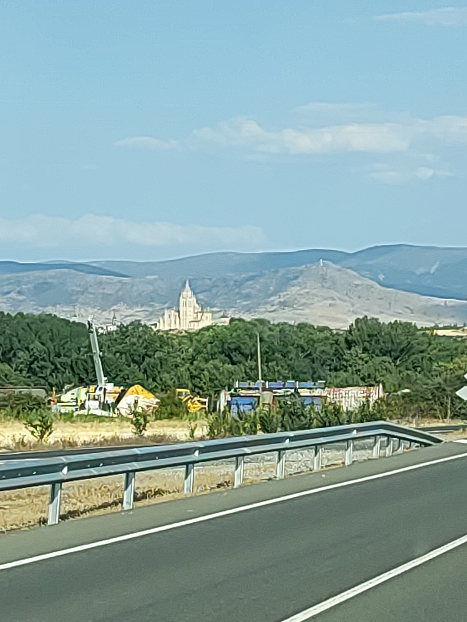 The image shows a panoramic view of Segovia, Spain, from a distance. In the foreground, a highway guardrail separates the road from a field with dry grass and sparse vegetation. Behind the guardrail, a dense green forest stretches across the mid-ground.
Prominently visible within the forest is the iconic Segovia Cathedral, a grand Gothic structure with a tall, multi-faceted spire. To the right of the cathedral, further in the distance and on a hill, stands a statue of Christ the King.
In the lower left portion of the mid-ground, amidst the trees, are various construction elements. A large crane, possibly yellow and white, is positioned near some assorted industrial equipment and debris, suggesting ongoing construction or a work site. The overall impression is a landscape that blends natural beauty with man-made structures, including historical landmarks and modern development. The sky is a clear blue with a few wispy clouds.