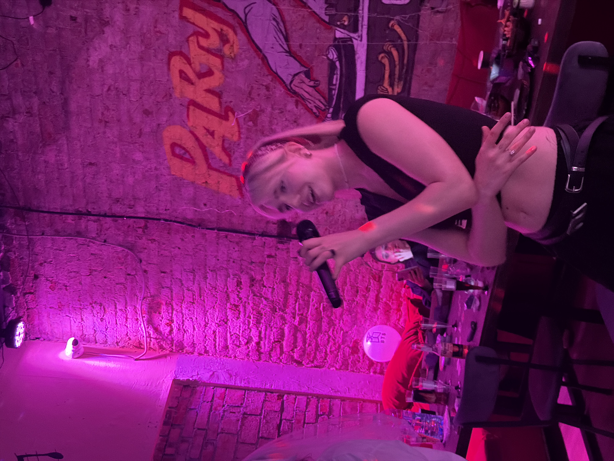 The image depicts a dimly lit, brick-walled room, likely a karaoke bar or party venue in Saint Petersburg, Russia, illuminated by vibrant pink lighting. A young woman with blonde hair, wearing a black crop top and a black skirt, is the central subject, holding a black microphone and appearing to sing. Her arms are crossed over her abdomen, and she is wearing a ring on her left hand. In the background, other individuals are visible, with one person seated at a table and another in the far background. The table is cluttered with glasses, bottles, and what appears to be food. A pink balloon with text is also present on the table. A graffiti-style mural with the word "Party" is painted on the brick wall behind the woman. The overall atmosphere suggests a lively social gathering.