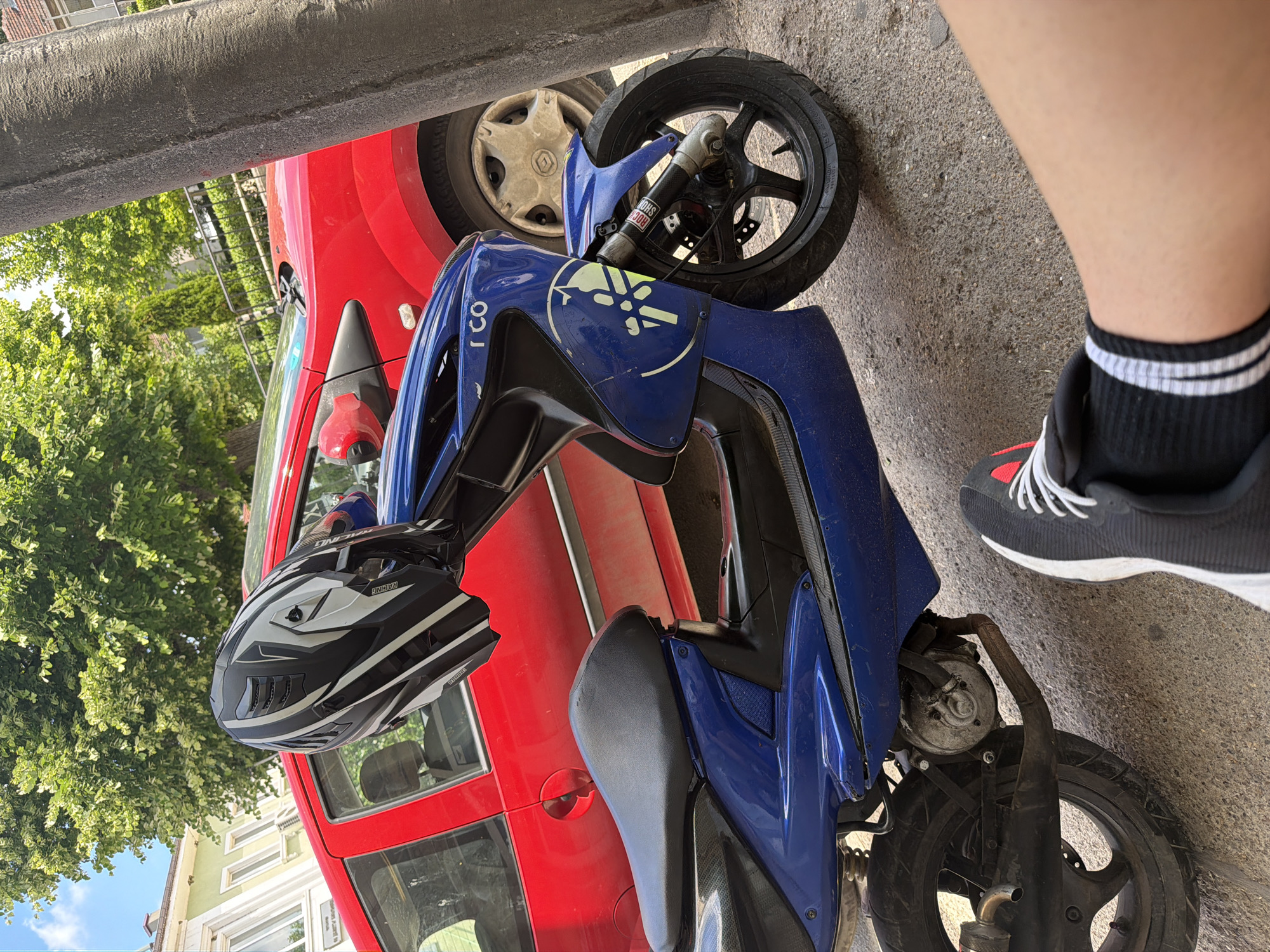 This image depicts an outdoor scene in Basarbovo, Bulgaria, under clear blue skies on a sunny day.
The primary subject is a blue Yamaha motor scooter, parked on an asphalt or concrete surface. A black, white, and grey motocross-style helmet, with "RACING" text visible, rests inverted on the scooter's handlebars. The scooter displays a white Yamaha logo graphic on its front fairing, alongside the text "I CO". A "ROCK SHOX" sticker is present on the front suspension fork. The exhaust system shows signs of corrosion.
Immediately behind the scooter is a red hatchback car, partially obscured on its right side by a textured grey concrete pillar. The car's front passenger side wheel, fitted with a light-colored hubcap, is visible. A noticeable dent or indentation is present on the driver's side rear door area.
In the foreground, the lower right leg, ankle, and foot of a person are visible, clad in a black sneaker with white laces and a black sock featuring white stripes.
The background features several lush green deciduous trees, indicating a warm season, and distant light-colored multi-story buildings with multiple windows. A black metal fence runs along the mid-ground behind the car.
