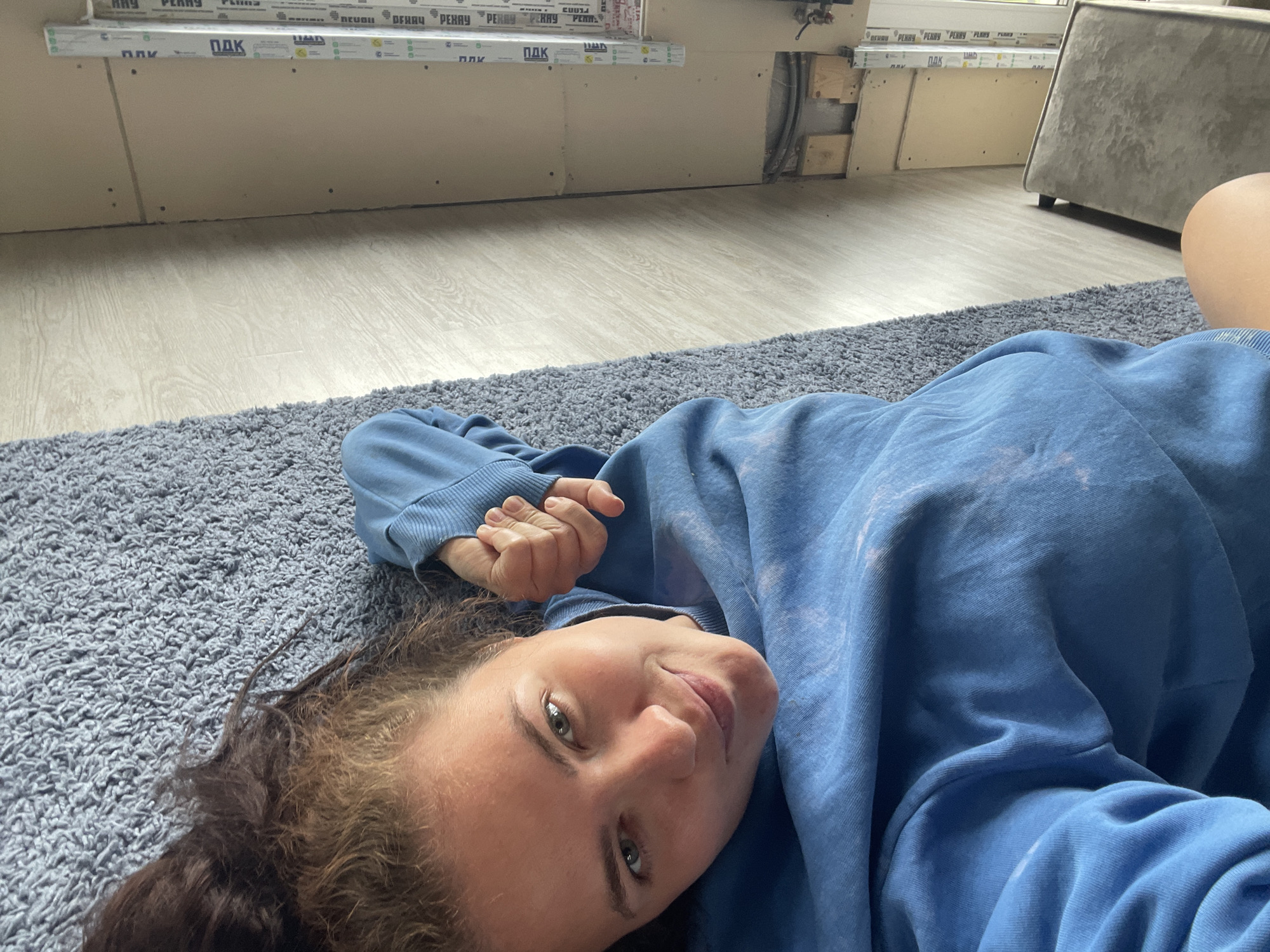A young woman with light skin, green eyes, and light brown hair lies on her side on a grey carpeted floor. She is wearing a blue hoodie. Her left arm is bent, and her hand is resting on her chest. The background shows a room under construction with drywall and some exposed pipes. A portion of a light-colored sofa is visible in the upper right corner. The lighting suggests an indoor setting during daytime. The location is described as Мосметаллоконструкция Онт, Russia.