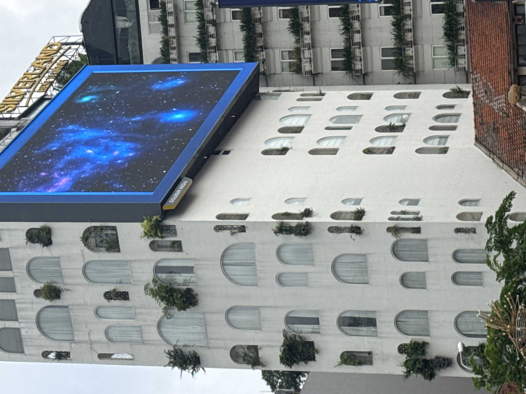 The image depicts a tall, white building in Thủ Đức, Vietnam, with a large LED billboard displaying a vibrant blue and purple nebula-like image. The building features a distinct architectural style with numerous arched windows, some of which are adorned with greenery. Small trees and plants are strategically placed on ledges and in niches on the building's facade, adding a touch of nature to the urban landscape. To the right of the white building, another gray building is visible, also featuring balconies with plants and windows. The sky is overcast. A sign reading "SILVERLAND BENTHANH" is visible atop a structure behind the billboard. The lower part of the image shows a red-tiled roof of a lower building, with some greenery in the foreground.