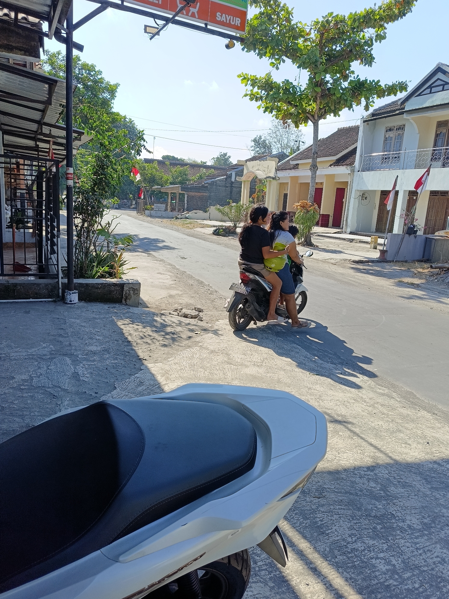 In the city of Borangan, Indonesia, two women ride a white scooter on a paved street. The woman in front wears a blue and white striped shirt and blue shorts, while the woman behind wears a black shirt and shorts. They are both looking forward. A green gas tank is visible between them, strapped to the scooter. In the foreground, the seat of another white scooter is visible. The street is lined with buildings on the right, and a sidewalk with a fence and vegetation on the left. Trees with green leaves are present, and the sky is clear and blue. Indonesian flags are flying in front of some buildings. The scene is bathed in sunlight, casting long shadows.
