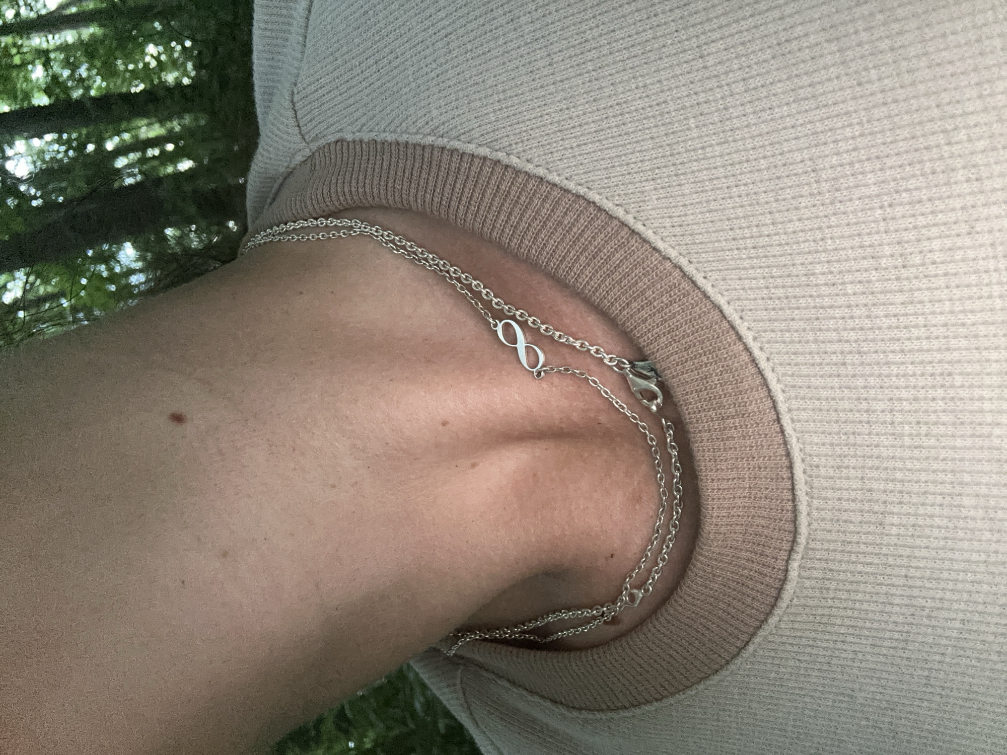 This is a close-up, eye-level shot of a person's neck and shoulder, wearing a light-colored, ribbed sweater. A delicate, silver necklace with an infinity symbol pendant rests on the person's skin, partially visible beneath the sweater's neckline. The necklace appears to have two separate chains, one slightly longer than the other, both adorned with small links. A silver clasp is visible near the top right of the frame. The background suggests an outdoor setting with dappled sunlight filtering through green foliage, hinting at trees and possibly a forest or park. The lighting is natural, suggesting daytime, and the overall mood is relaxed and casual. There is no visible text in the image.