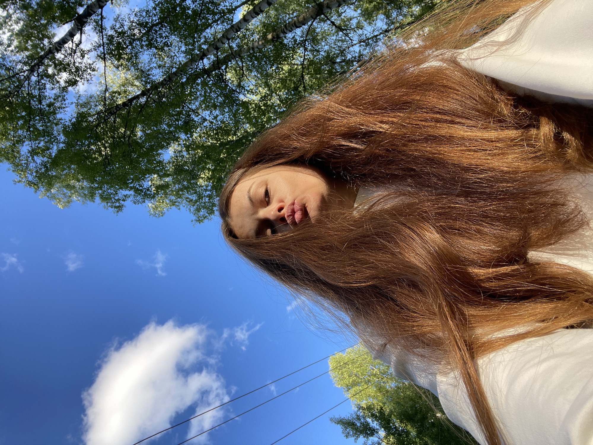 A young woman with long, auburn hair is captured in a low-angle, outdoor selfie. She is wearing a white sweatshirt and puckering her lips for the camera. Her face is partially obscured by her hair, which cascades around her. In the background, a vibrant blue sky is dotted with a fluffy white cloud on the left. Several dark power lines are visible against the sky. On the right side of the image, the leafy green canopy of trees, likely birch trees due to their distinctive white and black bark, forms a natural frame. The sunlight filters through the leaves, creating bright highlights. The scene appears to be set in a park or a wooded area within the city of Zarechny, Russia, likely during daylight hours.