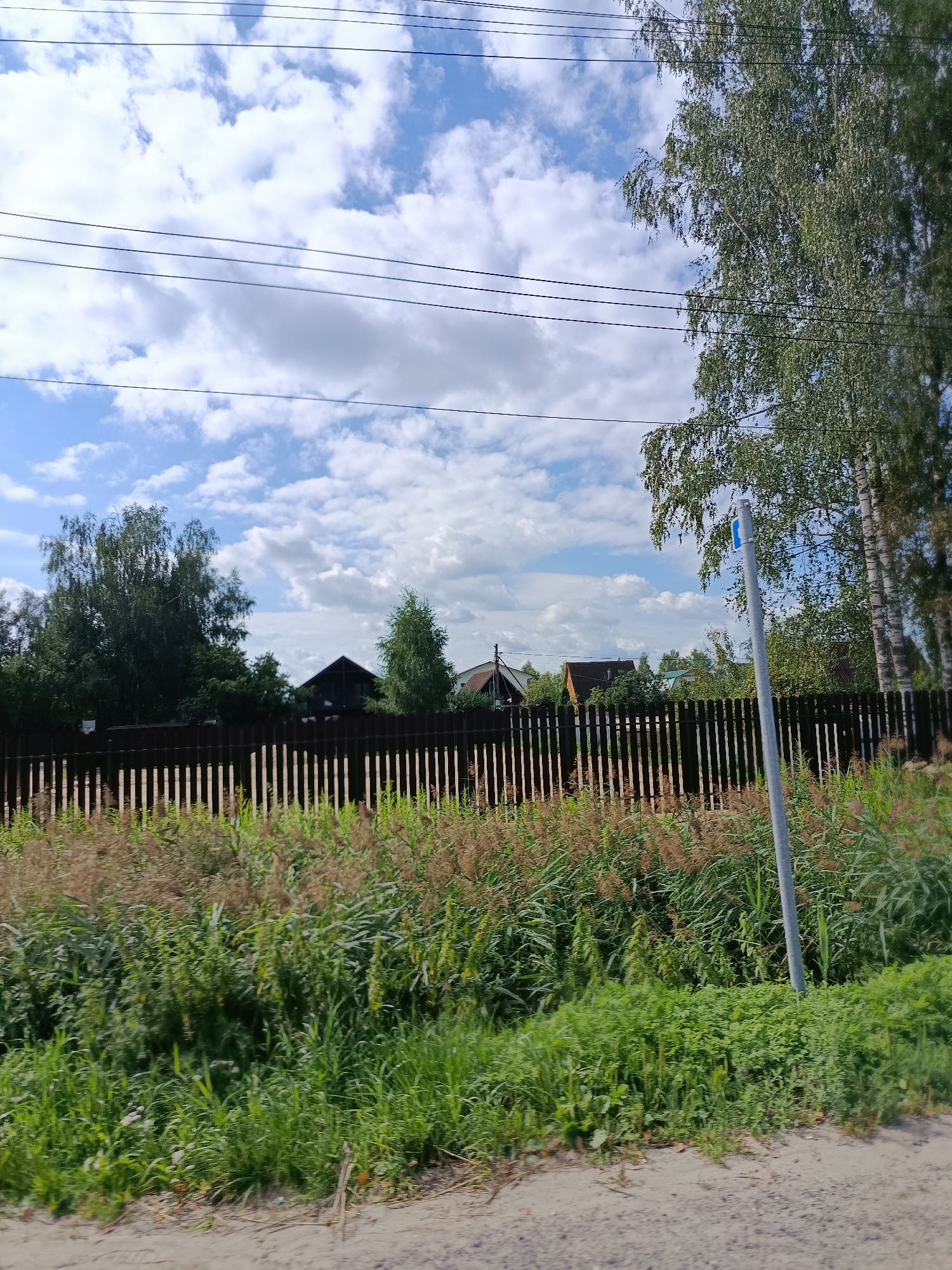 The image depicts a rural scene in Snopok Novyi, Russia, on a cloudy day. In the foreground, a dirt road is visible, bordered by lush green vegetation and taller, dry grasses. A dark brown wooden fence runs horizontally across the middle ground, separating the roadside from a cluster of houses and trees beyond. The houses are of varying sizes and designs, with some featuring dark roofs. Several large trees, including birch trees with their distinctive white bark, are scattered throughout the scene. Overhead, power lines stretch across the frame against a backdrop of blue sky and white, puffy clouds. A gray utility pole with a small blue sign is visible on the right side of the image, partially obscured by vegetation. The overall impression is of a quiet, residential area on the outskirts of a town.