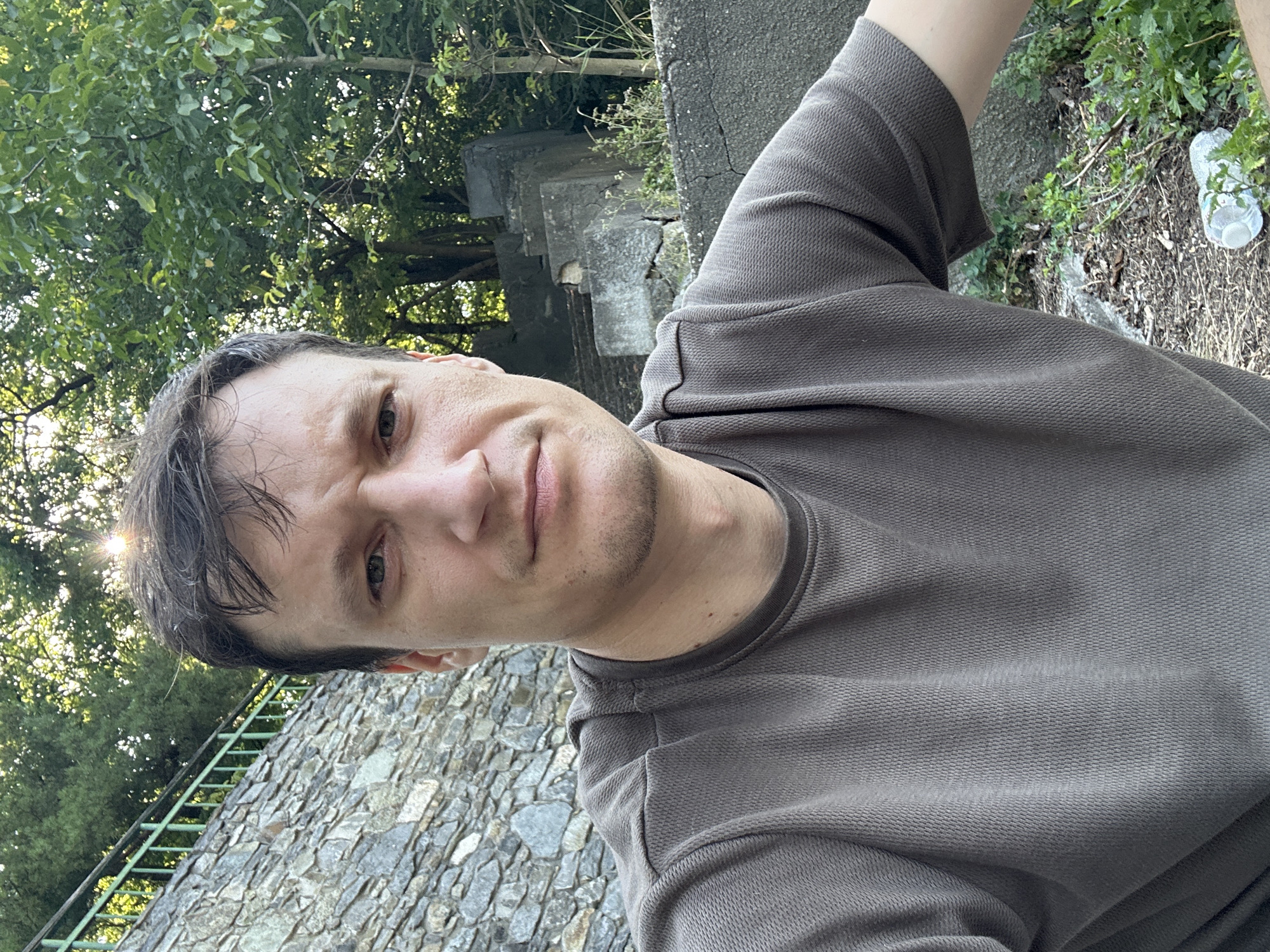 A medium shot captures a man with short, dark brown hair and a pensive expression. He is wearing a short-sleeved, textured, taupe shirt. The man is positioned outdoors, with a stone wall and green railing visible to his left, and lush green trees and foliage in the background. The sun is visible, peeking through the leaves of the trees. To the right, there are remnants of concrete structures and overgrown vegetation. On the ground in the lower right portion of the image, a plastic water bottle and some leaves are scattered. The overall scene suggests an outdoor, possibly rural or park-like setting within the city of Makiv, Ukraine.