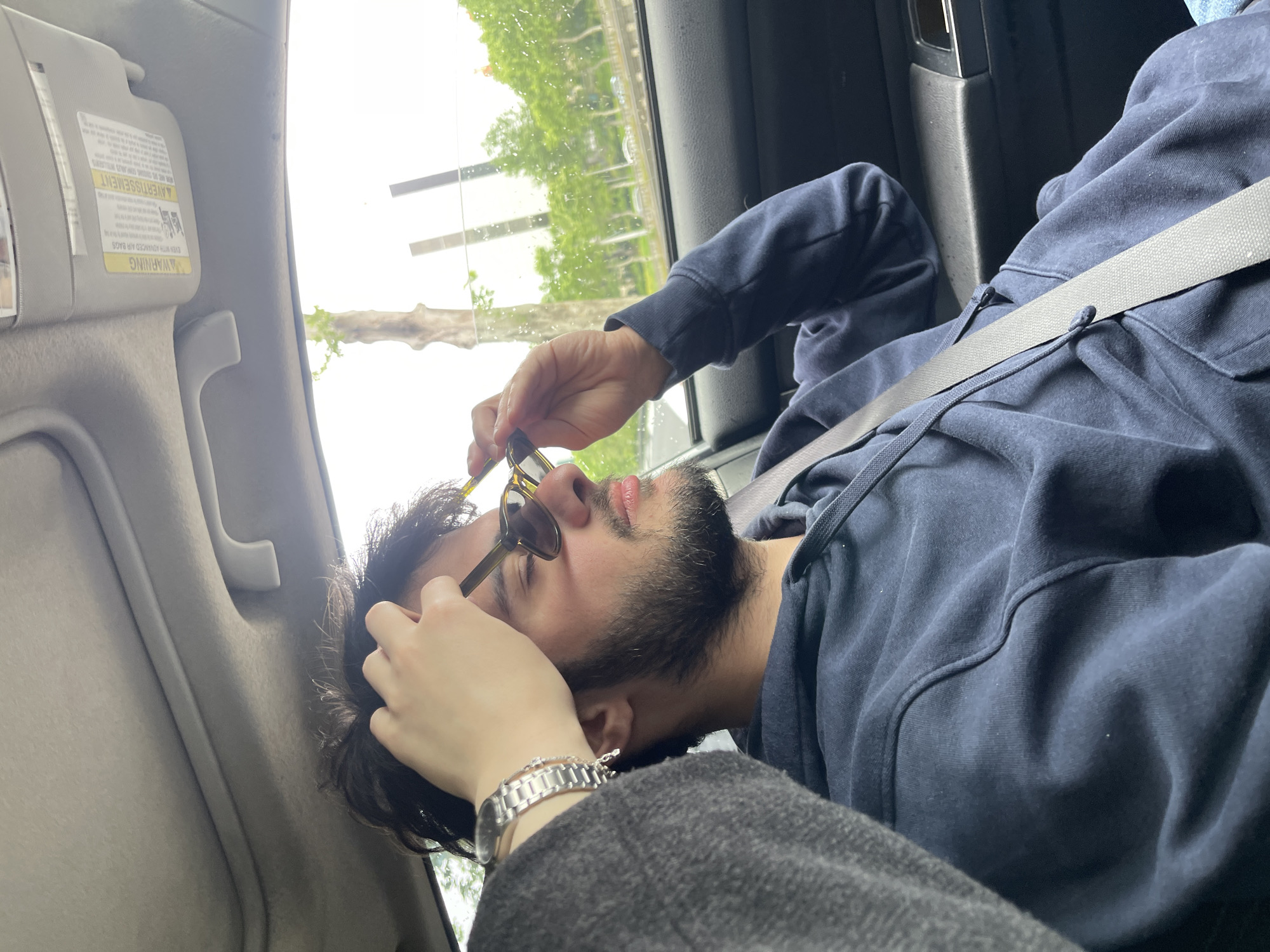 A person with a dark beard and sunglasses is seated in a car. They are wearing a navy blue hoodie and a silver wristwatch. The person is putting on their sunglasses with their right hand. The seatbelt is fastened across their chest. Outside the car window, trees and buildings are visible, suggesting an urban setting. The interior of the car includes a grey sun visor. The location could potentially be Tbilisi, Georgia based on building design and vegetation, but no specific landmarks are visible to confirm.