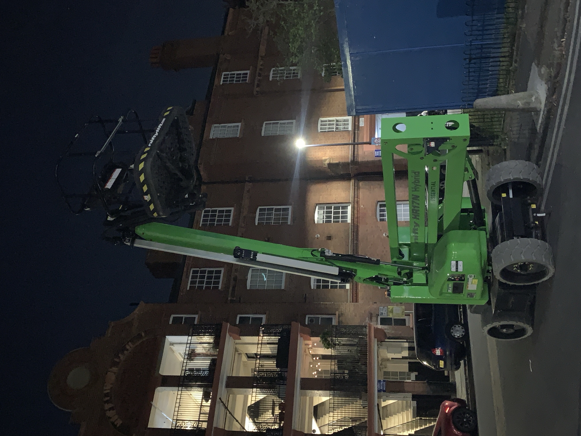 The image, taken at night or late evening, depicts an urban scene dominated by a large, bright green aerial work platform (AWP) against a backdrop of a brick building. The perspective suggests the camera is looking up at the AWP and the building.
The central subject is a Niftylift HR17N Hybrid boom lift, distinguished by its vibrant green color. Its extendable arm stretches upwards, culminating in a black work platform. This platform features prominent yellow and black diagonal striped safety markings, and the text "Niftylift.com" is visible on its side. Further branding on the main body of the lift reads "NIFTY HR17N Hybrid" and "ESSENTIAL". The lift appears stationary, suggesting it is either set up for a task or parked.
Behind the AWP stands a multi-story brick building with numerous white-framed windows, many of which are illuminated, indicating internal lighting. A bright street lamp shines prominently in the mid-ground, casting light on the building. To the right of the building, a large, dark blue barrier or structure is partially visible. The lower part of the frame reveals a paved surface, likely a street or sidewalk, along with a grey concrete bollard or barrier. Two vehicles are partially visible towards the bottom left: a dark blue car and the front section of a red car, suggesting a street parking or traffic scenario.
The sky is dark, confirming the nighttime setting. A few tree branches are visible near the top left, adding a natural element to the otherwise constructed environment. The scene is devoid of people, contributing to a quiet, utilitarian atmosphere. The architecture and street elements are consistent with an urban setting like London.