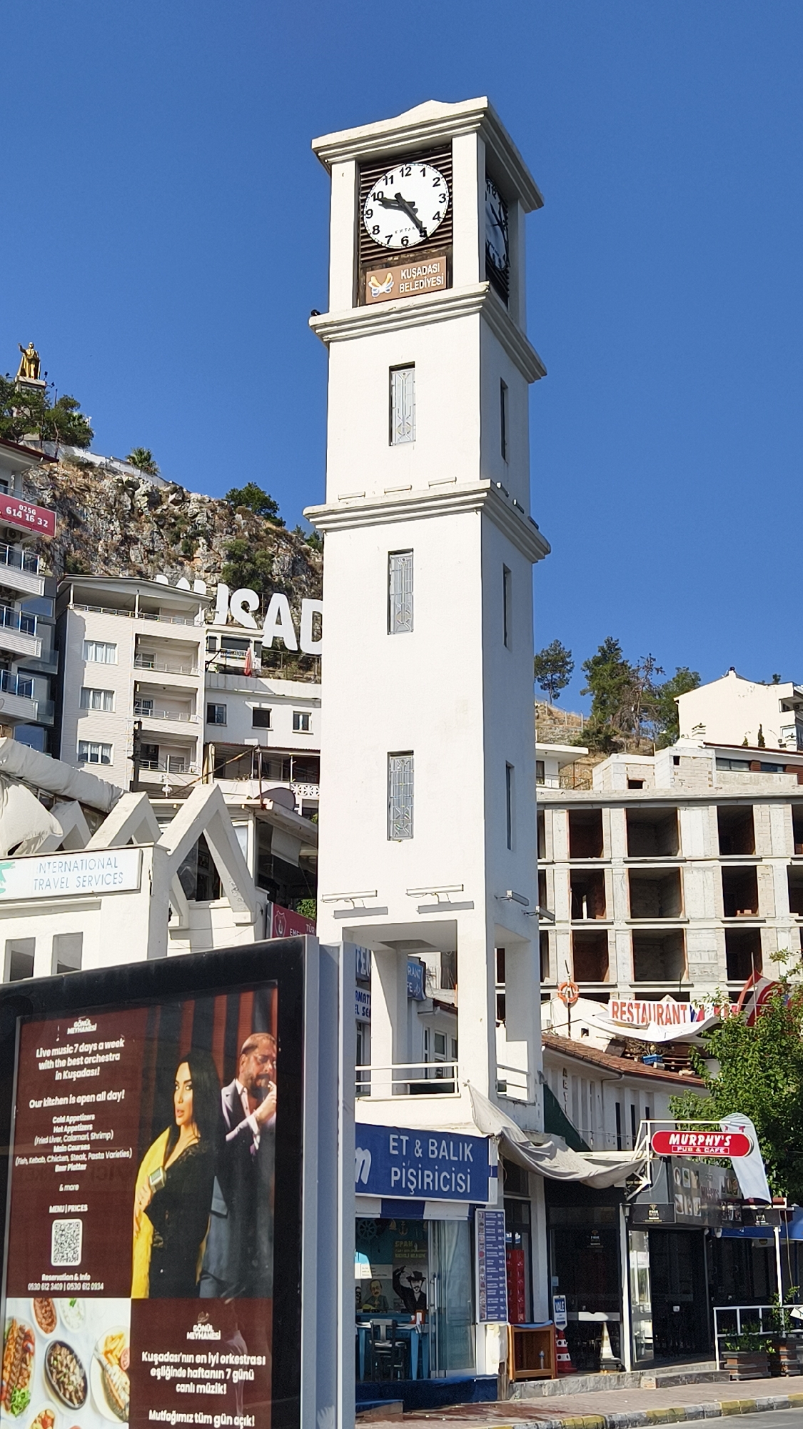 The image shows a prominent white clock tower in the city of Kuşadası, Turkey, against a clear blue sky. The clock tower features a large clock face with Roman numerals, and below it, a plaque with the text "KUŞADASI BELEDİYESİ" (Kuşadası Municipality). Buildings of various heights and architectural styles are visible behind and around the clock tower, including apartment buildings and structures under construction. The foreground displays a large digital billboard with an advertisement featuring two individuals and text in Turkish, along with a QR code and contact information. Further to the right, storefronts for businesses such as "ET & BALIK PİŞİRİCİSİ" (Fish and Meat Cookery) and "MURPHY'S PUB & CAFE" are visible. A statue of a figure is perched on a hill in the background, overlooking the city. The overall scene depicts a bustling urban environment with a blend of commercial and residential structures.