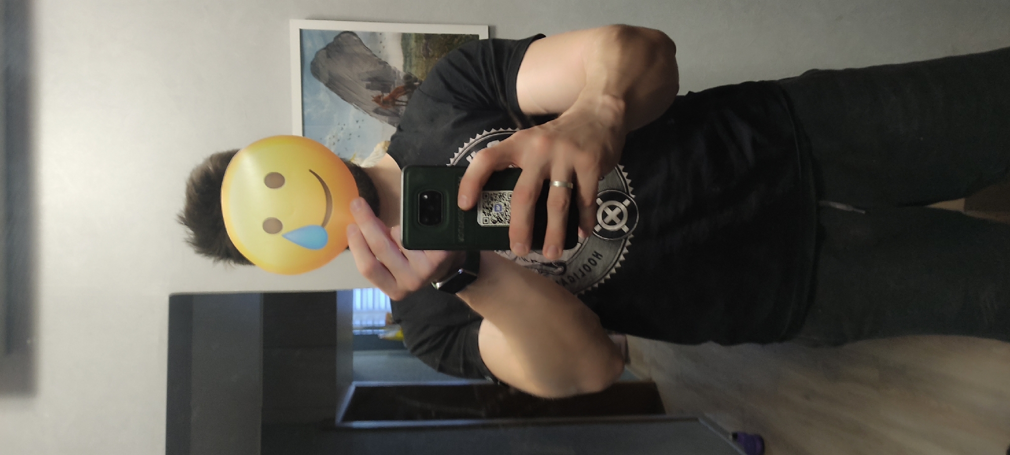 The image shows a man in a mirror selfie. The man has a yellow crying laughing emoji sticker over his face. He is wearing a black t-shirt with a design on it. He is also wearing a wedding ring and an Apple Watch. He is holding a green smartphone. Behind him is a framed picture on the wall. The room seems to be an apartment. It is not possible to determine if the image was taken in Fryazino, Russia.