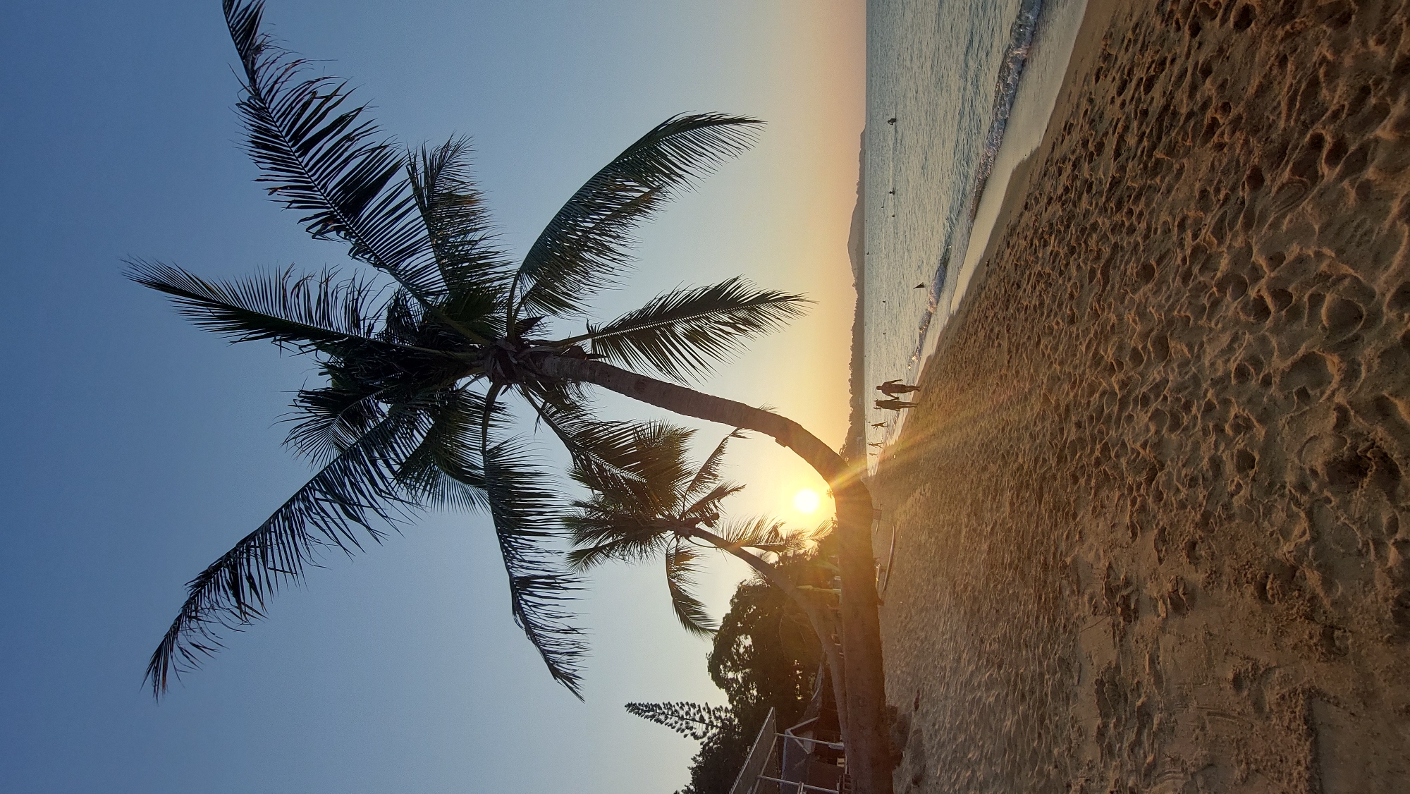 The image captures a sunset scene on a beach in Baan Mae Nam, Thailand. Two palm trees are prominently featured, their silhouettes set against the backdrop of the setting sun. The sun is positioned low on the horizon, casting a golden light across the sand and water. Several people can be seen walking along the shoreline in the distance. The sand shows numerous footprints, and the waves are gently lapping at the shore. The sky transitions from a clear blue to a gradient of orange and yellow near the horizon. Some vegetation is visible in the lower right, next to the trunk of the one of the palm trees.