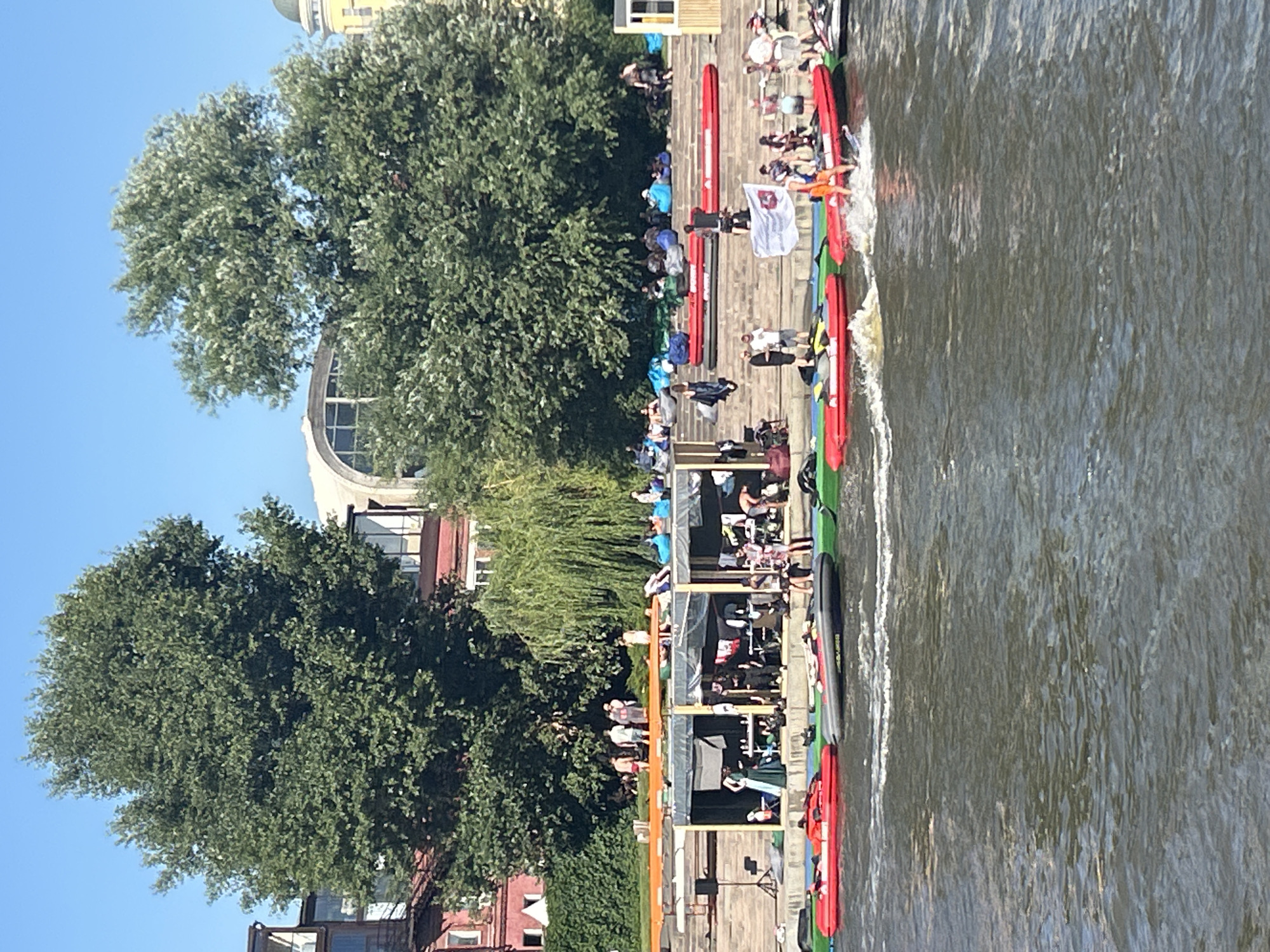 The image depicts a sunny day at a waterfront recreational area in Moscow, Russia, likely on the Moskva River embankment. In the foreground, several inflatable boats, predominantly red and gray, are present on the water. One person is standing in a red inflatable boat, appearing to be on a personal watercraft, creating a splash. The middle ground shows a tiered embankment with people relaxing on beanbag chairs and lounging on a platform. There are also structures resembling outdoor cafes or seating areas with tables and what appear to be some goods or supplies. Further in the background, large trees, one with distinctive weeping branches, and buildings, including a multi-story red brick structure and a building with a large arched window, are visible against a clear blue sky. People are seen walking and gathering on the embankment. The overall scene suggests a lively, outdoor leisure activity in a cityscape.