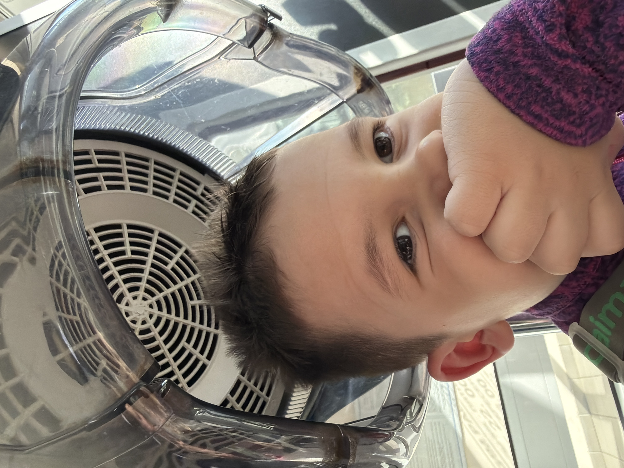 A close-up, slightly tilted image shows a young child's face pressed against a transparent plastic-encased white circular grille, presumably part of a mechanical device. The child's right hand is raised to their face, covering part of their mouth and cheek. The child has dark hair, brown eyes, and fair skin. They are wearing a purple patterned sweater. Light reflects off the plastic and grille, creating highlights. The location is unknown.