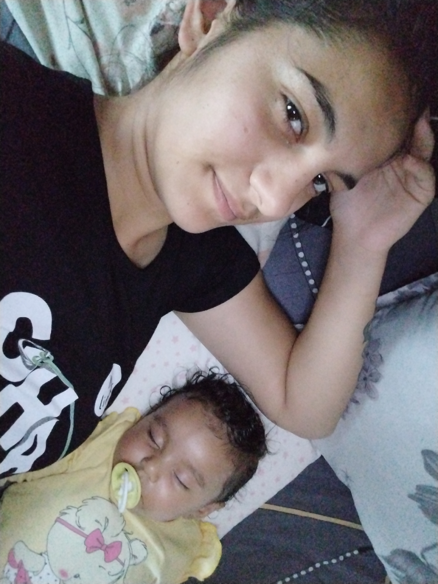A woman with dark hair, wearing a black t-shirt with white letters, lies in bed next to an infant in a yellow onesie with a pacifier in its mouth. They are lying on a bed with various patterned sheets and pillows.