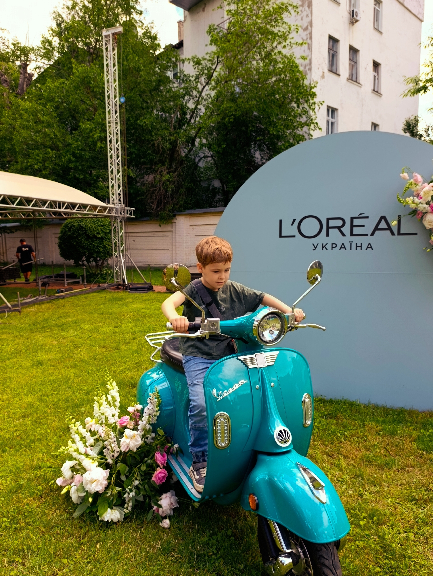 A young boy with blond hair sits on a turquoise Vespa scooter in a grassy outdoor area. He is wearing a dark green t-shirt and blue jeans. The scooter is decorated with a large bouquet of pink and white flowers at its base. Behind the scooter, a large, light blue circular sign displays the L'Oréal logo and the text "УКРАЇНА" (Ukraine) in black. The backdrop includes a white building with windows and trees. In the background to the left, a man in a dark t-shirt is visible near some event equipment and a white tent structure. The lighting suggests it is daytime. The scene appears to be set up for an outdoor event, possibly a promotional event in Kyiv, Ukraine.