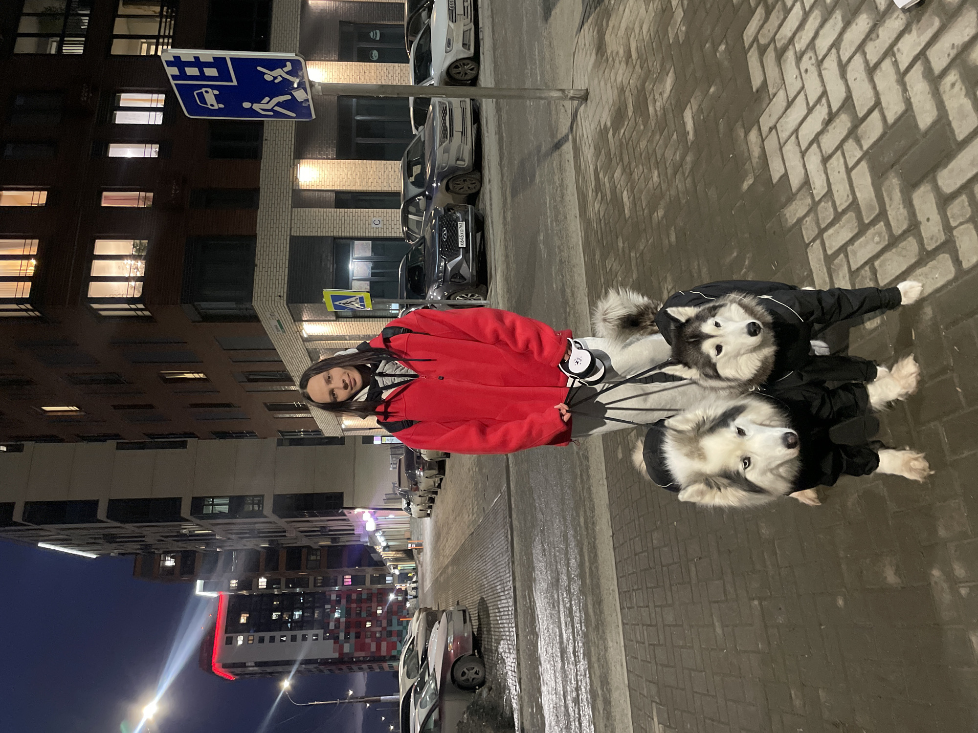 The image depicts a woman and two husky dogs on a wet, brick-paved street in what appears to be an urban environment, possibly Izhevsk, Russia, based on available information. The woman, wearing a red jacket and light-colored pants, stands near the center of the frame. The two dogs are next to her and appear to be wearing black jackets. The background includes a variety of buildings, including residential structures with illuminated windows. Cars are parked alongside the street. A street sign is visible overhead. The overall scene is illuminated by streetlights and building lights, indicating it is evening or night.