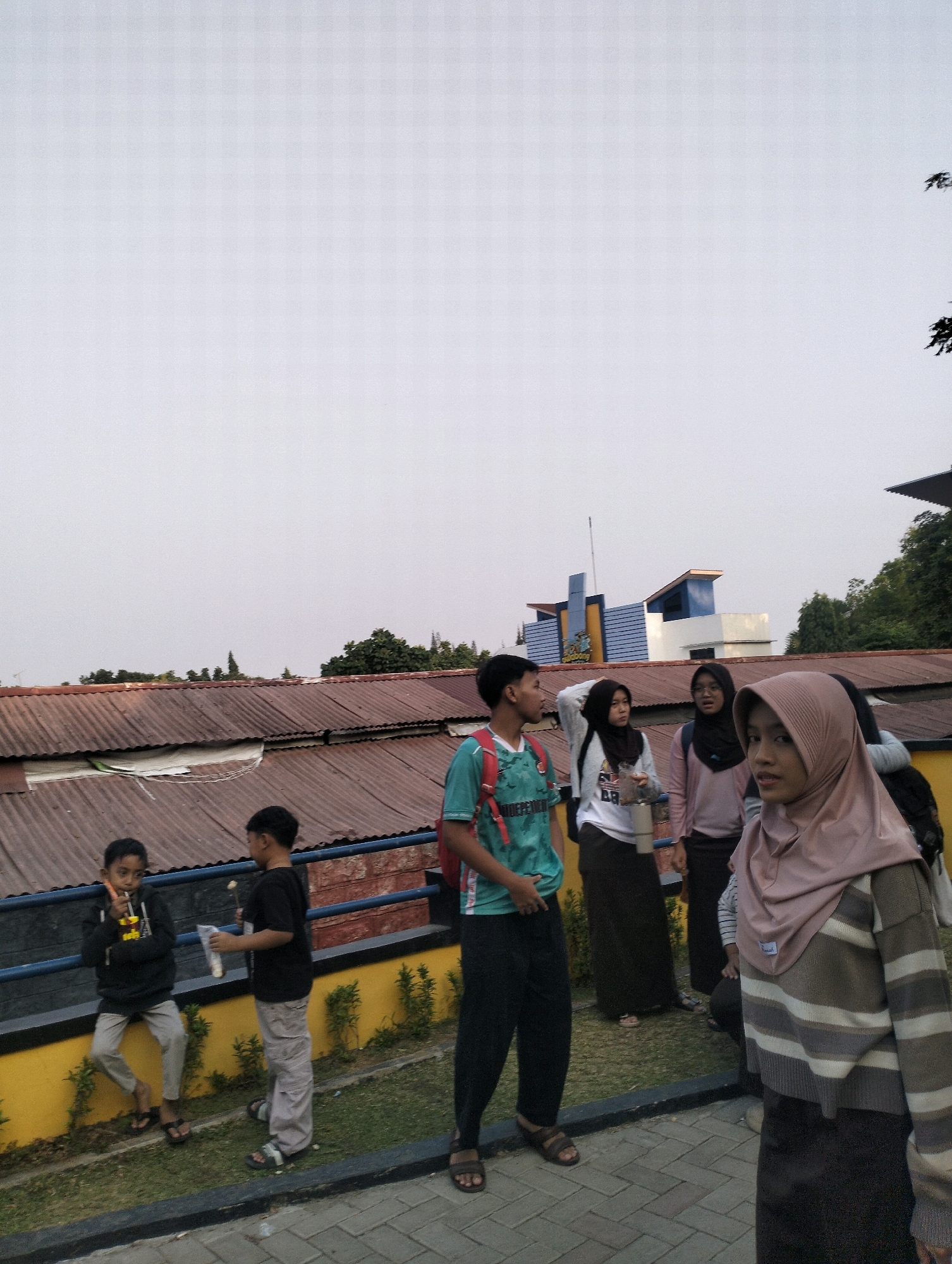 In Karangbanjar, Indonesia, a group of individuals is gathered outdoors. In the foreground, a young person wearing a light brown and grey striped sweater and a dark skirt is facing the camera, while a person with a pink hijab stands nearby. To the left, a young boy in a black hoodie and grey pants holds a cup and straw, and another boy in a black t-shirt and grey pants stands with a plastic cup. Further back, a young person in a green t-shirt and black pants stands with a backpack, alongside two young women in hijabs and long skirts, one of whom is holding a white cup. The background features buildings with corrugated metal roofs and some vegetation. The sky is overcast.