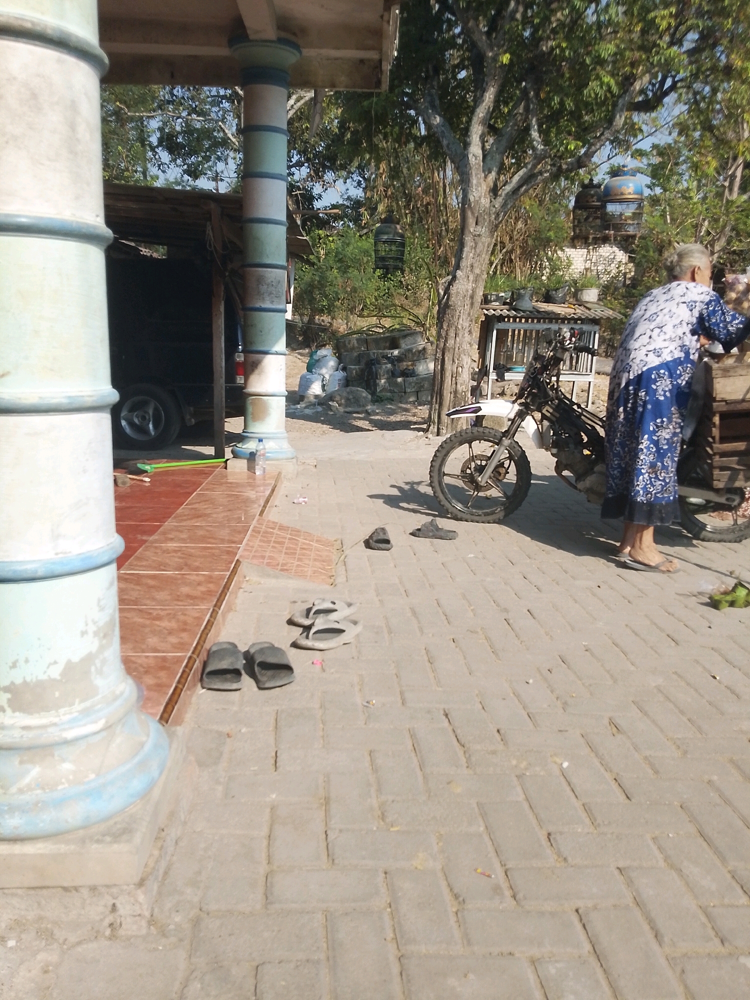 A person wearing a blue and white floral-patterned dress stands near a motorcycle parked on a paved area in Trenggulunan, Indonesia. The person's feet are visible, wearing flip-flops. A white motorcycle with a dirt bike-style tire is positioned on a paved surface. Several pairs of flip-flops are scattered on the pavement in the foreground and middle ground. In the background, there are trees and structures, including a wooden shelter with a corrugated metal roof. A large white pillar is visible on the left side of the image, and another pillar with alternating bands of white, green, and blue is situated in the middle ground. A car tire is visible inside a dark opening to the left of the pillar. The ground is covered with interlocking brick pavers. The scene appears to be outdoors, under daylight.
